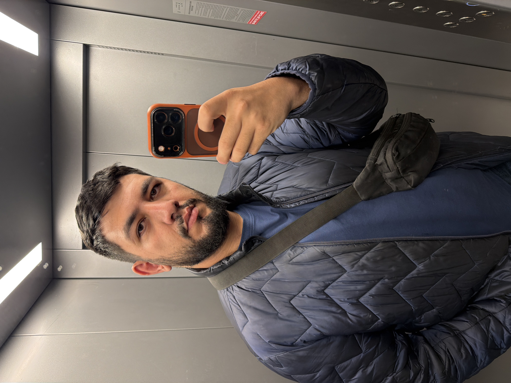 A man is taking a selfie in an elevator with a phone in an orange case. He has dark hair and a beard, and is wearing a navy blue quilted jacket with a crossbody bag. The elevator has gray walls and ceiling with recessed lighting. The location is Krasnoyarsk, Russia.