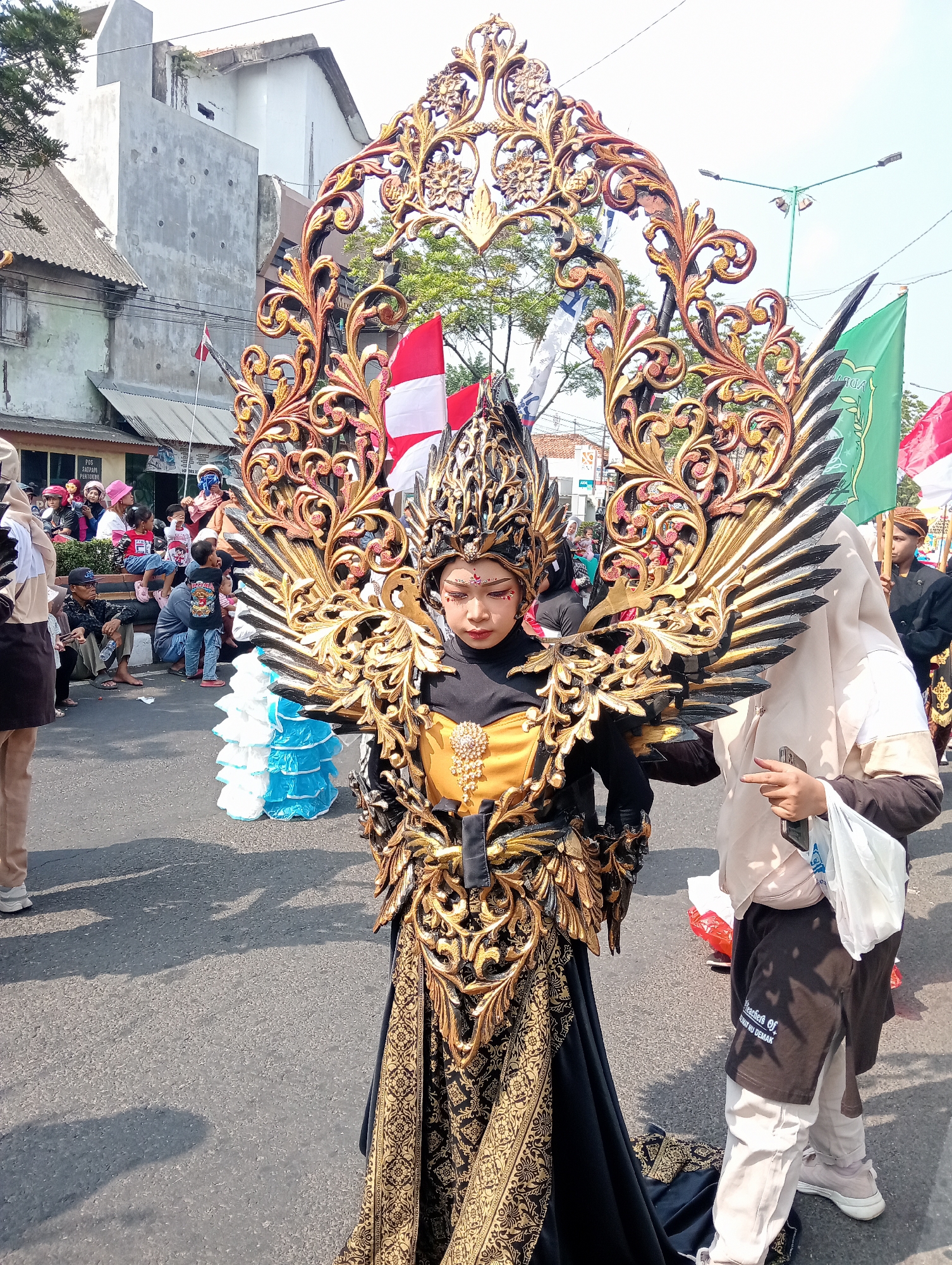 In the city of Bintoro, Indonesia, a person in an elaborate golden costume, featuring wings and a headdress resembling a crown, stands in the middle of a street. The costume is adorned with intricate patterns and appears to be made of gilded material. The person's face is partially visible, and they are wearing makeup, including red accents around the eyes and on the cheeks. Behind this central figure, spectators are gathered, some sitting on the roadside, and others standing. In the background, buildings are visible, along with flags, including Indonesian flags and a green flag. The scene suggests a parade or cultural event taking place on a sunny day.