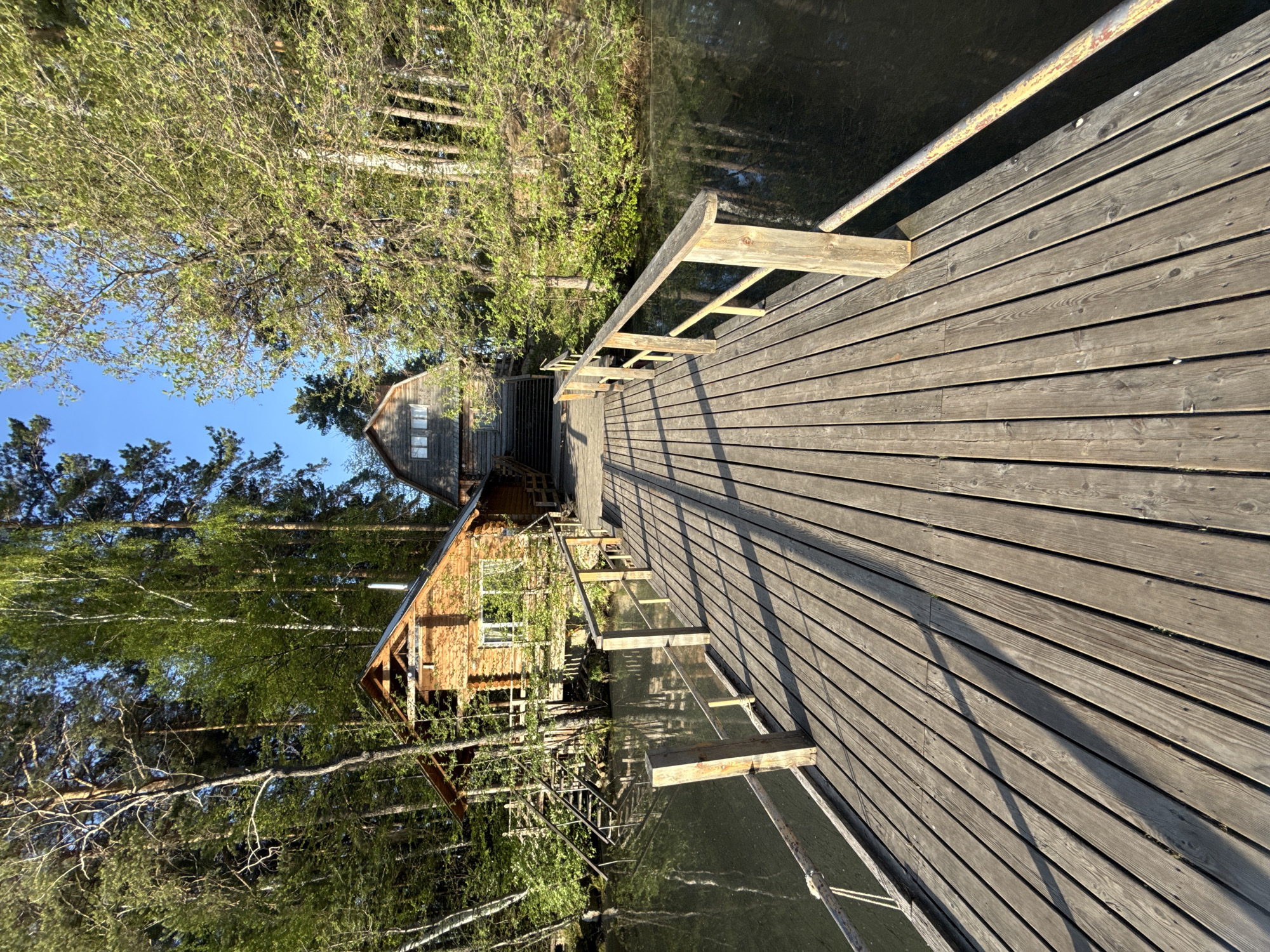 A wooden pier extends over water towards a wooden cabin in Zolotoi Plyazh, Russia. The pier is made of wooden planks, with a metal railing visible along one side. Wooden frames and railings appear along the edge of the pier. The cabin is constructed of wood and features windows. Trees surround the cabin and line the opposite side of the pier. The water appears dark and reflective. The sky is visible through the trees and is blue.