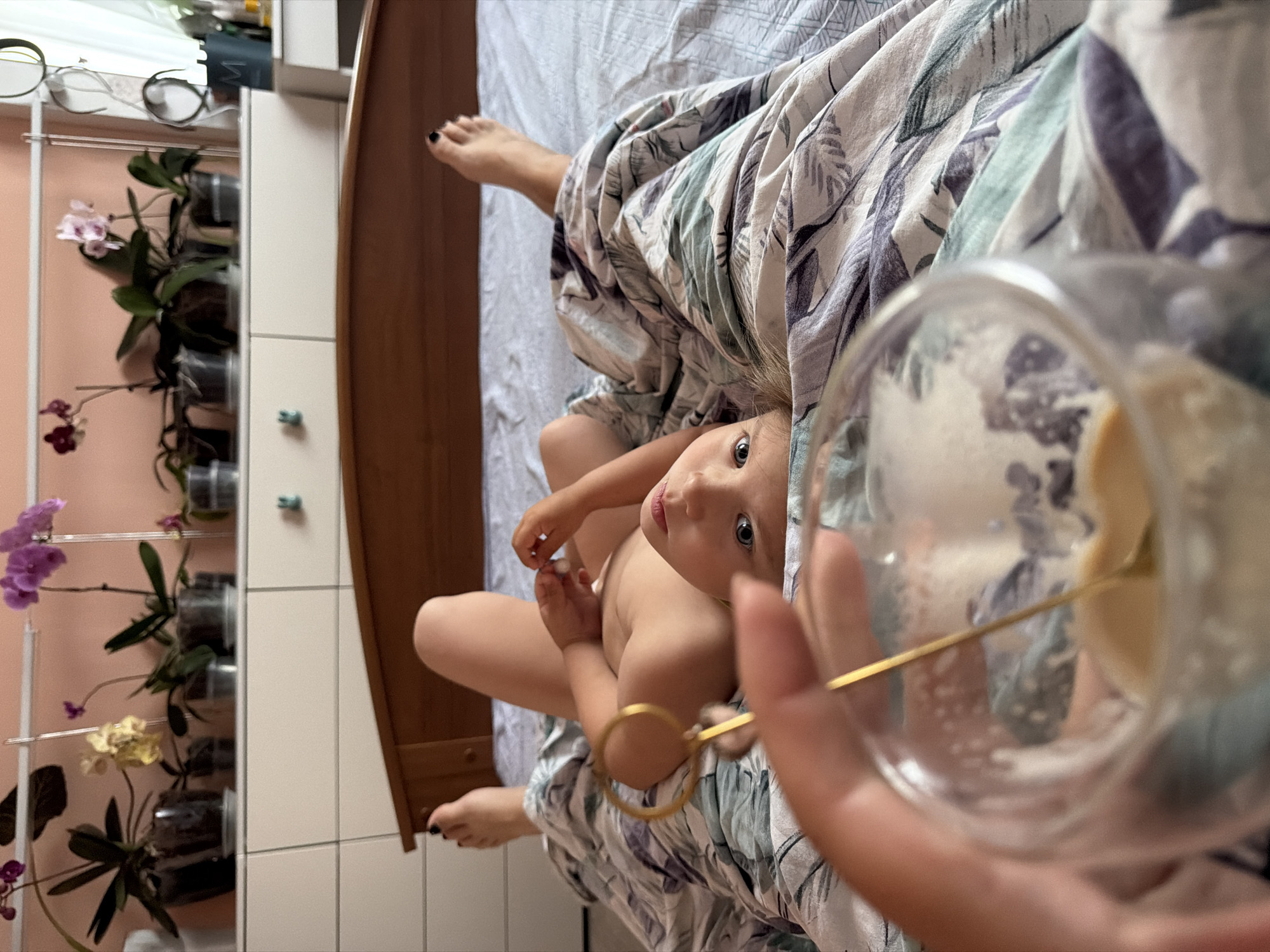 A child lies on a bed under a floral patterned blanket, gazing upwards. A hand holds a glass with a golden spoon inside, partially obscuring the view. To the left is a white cabinet with potted orchids on shelves. The setting appears to be indoors. The location is not identifiable based on the visual information provided.