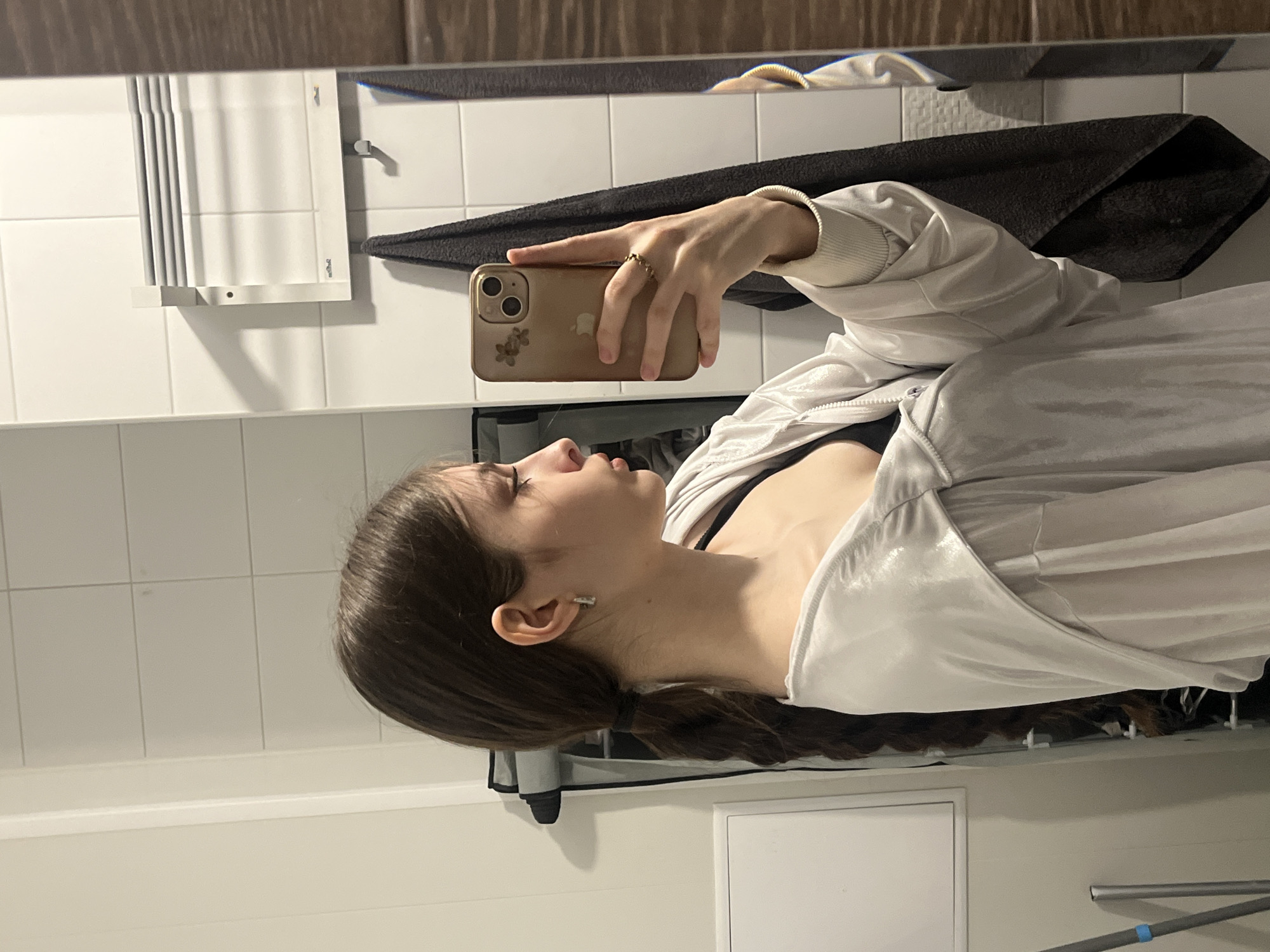 The image captures a mirror selfie of a young female, positioned horizontally as if she is looking to her right. She has long, dark brown hair styled in a thick braid that rests over her shoulder. She is wearing a light-colored, possibly white or silver, shiny zippered jacket over a black top or bra. A small, gold-colored ring with a beaded design is visible on the ring finger of her right hand, which holds a light gold-colored iPhone with a clear case featuring a delicate floral pattern. A small, silver-colored earring is visible in her left ear.
The setting appears to be a bathroom, evidenced by the white rectangular wall tiles that cover most of the background. A dark grey or black towel hangs on a rail behind her arm. To her left, a white panel radiator or heater is mounted on the wall. The top edge of the mirror reflects a dark wooden surface, possibly a cabinet or ceiling trim. Below the radiator, a white rack is visible. The activity taking place is the subject taking her own photo using a mirror. The lighting is artificial, suggesting an indoor environment. No discernible text is visible in the image.