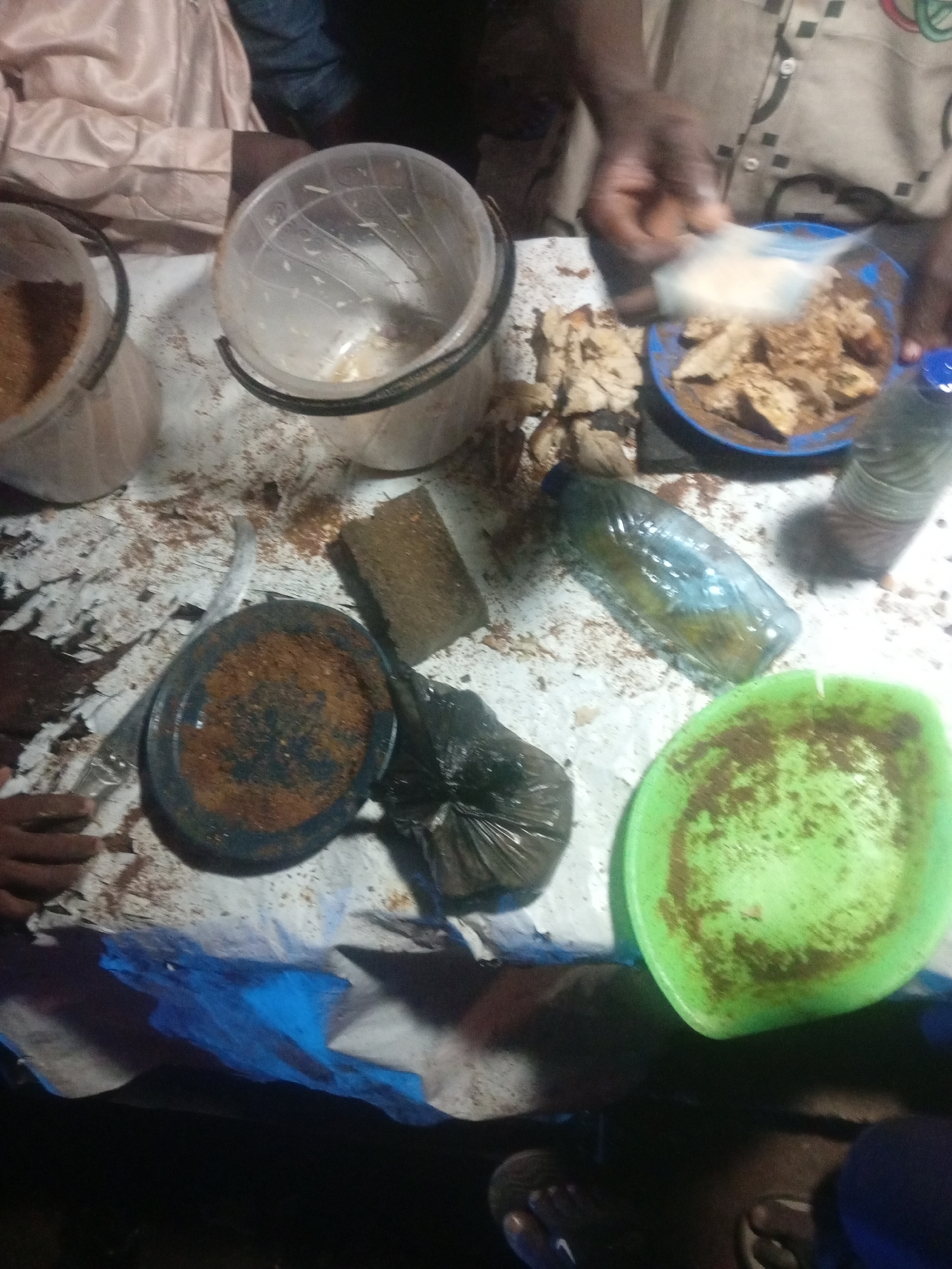 In an outdoor setting, likely in Tundun Wada West, Nigeria, a group of individuals are engaged in preparing food, possibly for a communal meal or a street vending operation. The scene is cluttered with various food preparation items. Two translucent plastic buckets are visible, one containing what appears to be finely ground grain or flour, and the other empty and slightly grimy. A blue bowl holds pieces of cooked food, possibly yam or cassava, which are being seasoned. A person's hands are adding a white powdery substance, likely spices or seasonings, to the bowl.
Other objects include a plastic water bottle, partially filled with a yellowish liquid, and a black plastic bag containing more of the cooked food or ingredients. A dark-colored plate with a reddish-brown residue, likely spices, sits beside a sponge-like object and a piece of brown, dried material. A bright green plastic plate, also with residue, is positioned towards the foreground.
The ground beneath the makeshift table appears to be dirt, with some blue plastic sheeting partially covering the area, suggesting an informal or temporary setup. The lighting suggests it is daytime, though the exact time is difficult to determine. The overall impression is one of active food preparation in a rural or semi-urban environment. No discernible text is present in the image.