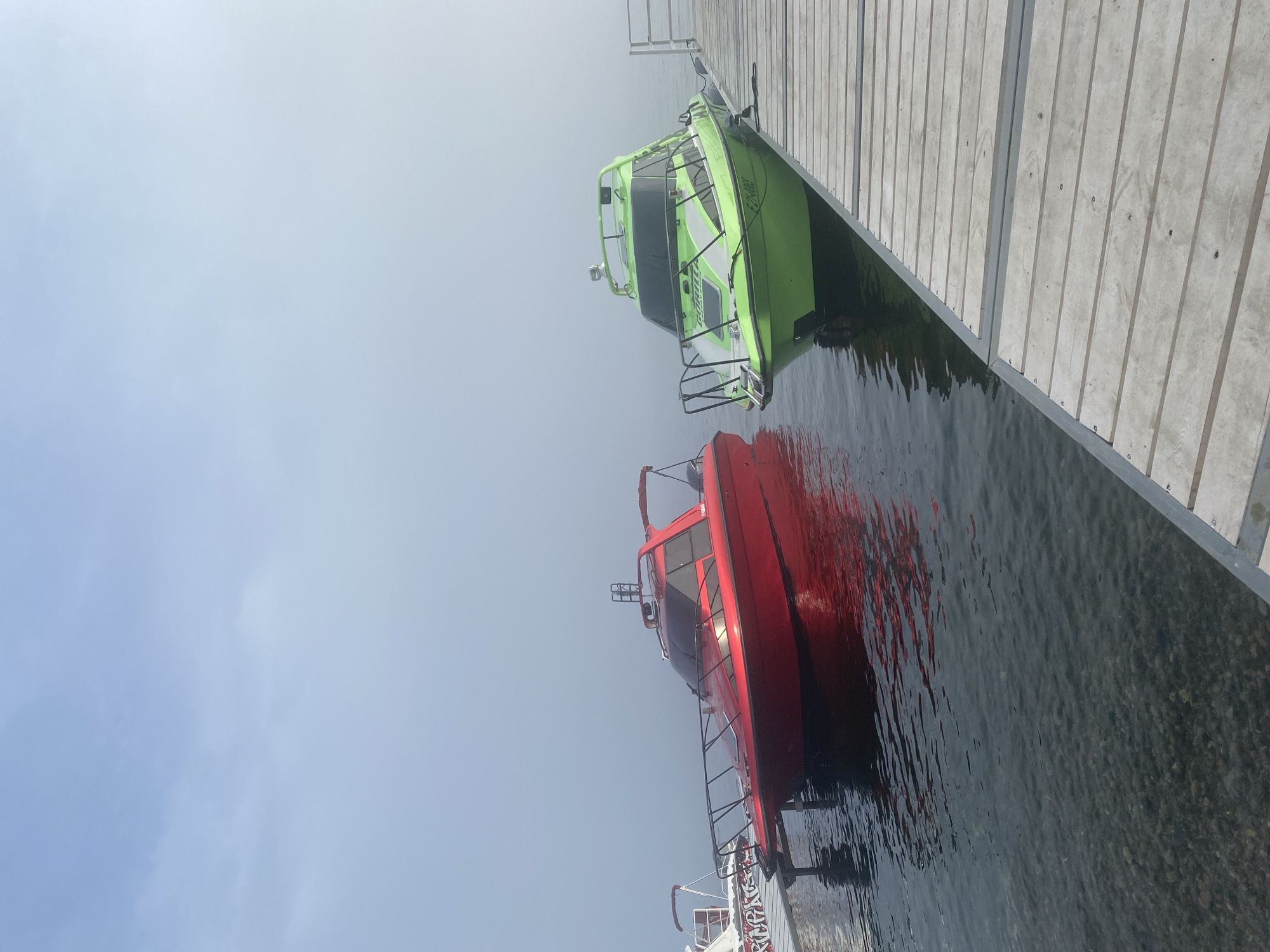 Two motorboats, one red and one lime green, are docked at a wooden pier on a body of water. The boats are modern speedboats with enclosed cabins and railings. The red boat is positioned to the left, and the green boat to the right, closer to the camera. The water is calm and shows a clear view of the pebbles on the lakebed. The background is obscured by fog, with the sky a pale blue. A portion of another white boat with graffiti-like lettering is visible to the far left. The scene appears to be a foggy morning in Listvyanka, Russia, a settlement on the southwestern shore of Lake Baikal.