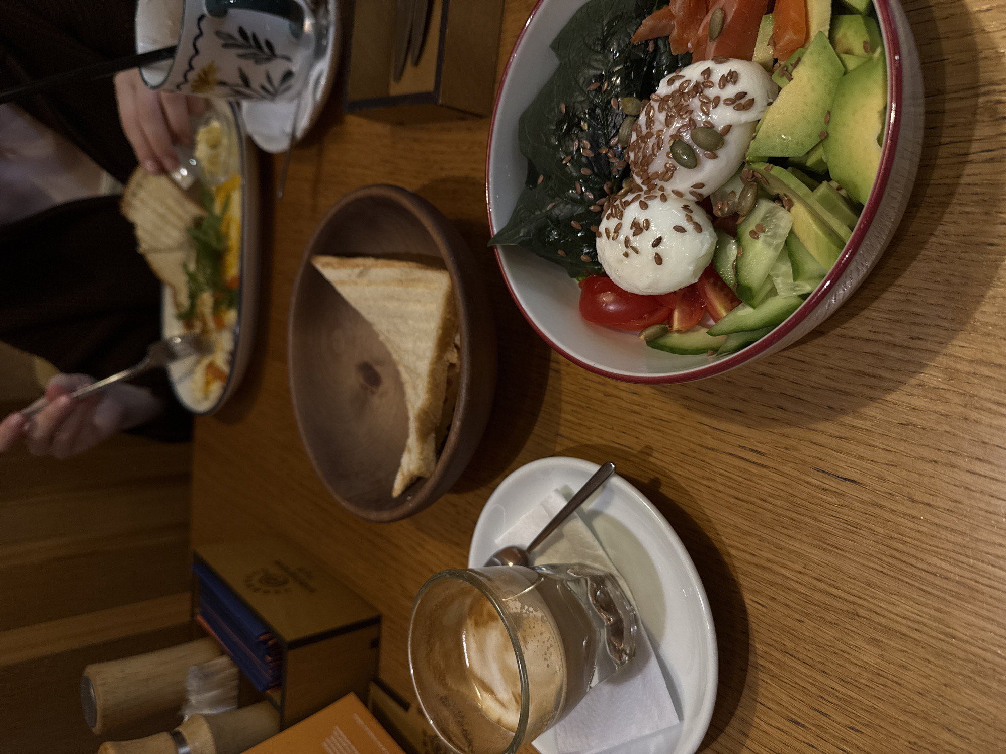 The image shows a table setting with a bowl of salad containing greens, tomatoes, avocado, mozzarella, and seeds. There's also a bowl of toast, a plate with what appears to be eggs and vegetables, and a glass of coffee with a spoon resting on a saucer. The items are placed on a wooden table. The setting is indoors and depicts a meal being consumed. Based on the details visible in the picture, it's difficult to precisely determine the location in Krasnoyarsk.
