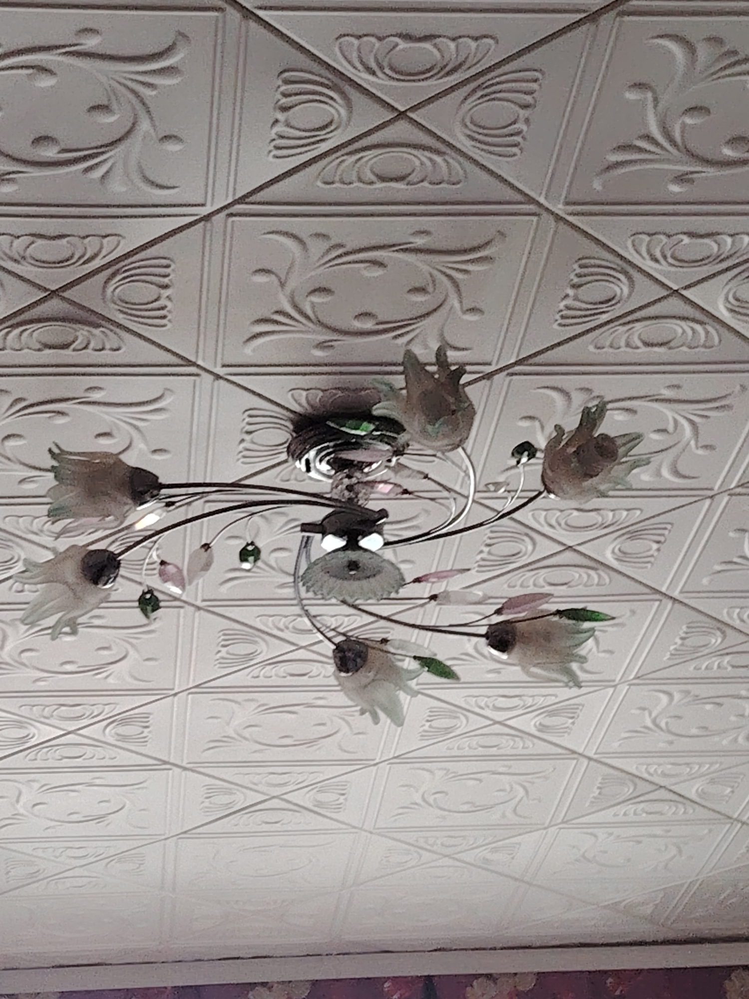 The media file displays an upward-facing view of an indoor ceiling in Yanaul, Russia, featuring a decorative chandelier against a patterned ceiling. The ceiling is covered with numerous light-colored, possibly white or off-white, square tiles. These tiles are embossed with intricate designs, predominantly a repeating grid of squares. Within these squares, two main patterns alternate: one is a graceful, scrolling floral or vine motif, and the other is a more geometric, concentric, or shell-like design. Diagonal lines run across the ceiling, sometimes intersecting the corners of the square tiles, adding another layer to the pattern.
A prominent chandelier hangs from the center of the ceiling. It features multiple curved, dark-colored metal arms emanating from a central metallic base. Each arm holds a translucent, light-colored shade, shaped like an inverted bell or a tulip, resembling frosted glass or plastic flowers. Dainty, leaf-shaped decorative beads or pendants, some green and some light pink or purple, are interspersed among the arms, adding splashes of color.
At the very bottom of the image, a narrow strip of a wall is visible, showing what appears to be a patterned wallpaper or border with floral designs, further indicating an indoor setting. There are no people visible, and no specific activity is taking place. The image is a static shot of the room's decor. No text is visible in the frame.