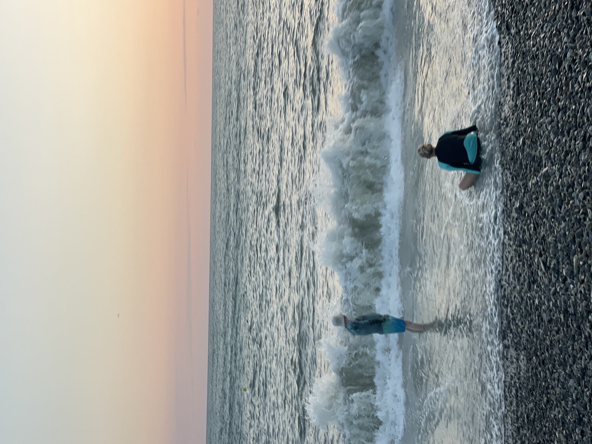 The image depicts a scene at a pebble beach in Sochi, Russia, at dusk. The sky displays a gradient of soft pastel colors, transitioning from pale yellow at the top to pink and orange near the horizon. The sea is moderately wavy, with a significant wave breaking near the shore. Two individuals are present: one person, appearing to be a child, stands in the shallow water wearing a hat and swim trunks, facing the sea. Another person, an adult, sits in the water facing away from the camera, wearing a rash guard with black and turquoise colors. The beach is covered with dark, rounded pebbles. The overall atmosphere is tranquil and serene, characteristic of a peaceful evening by the Black Sea.