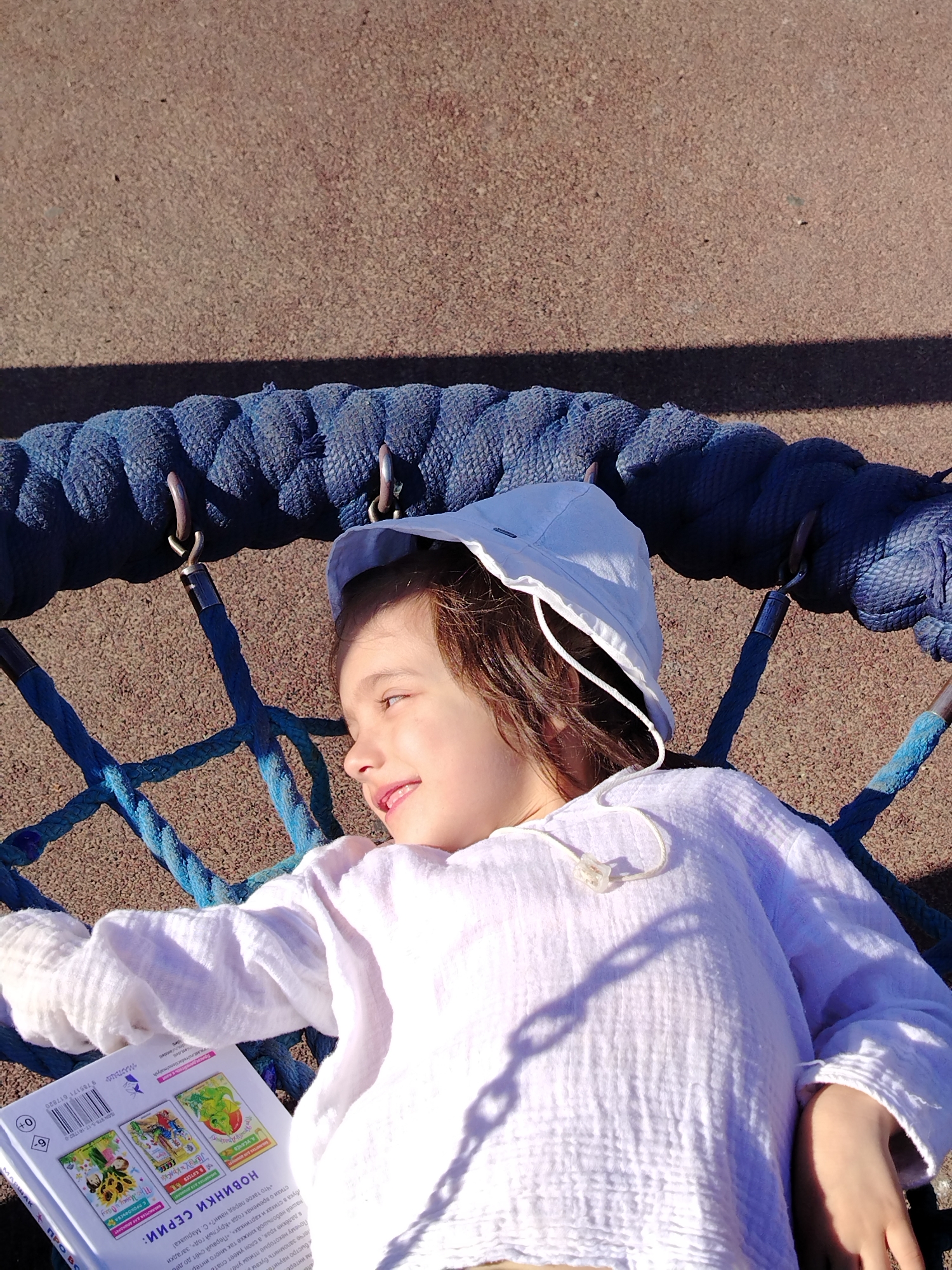 A young child is lying in a blue net swing. They are wearing a white shirt and a white sun hat. The child is holding a book titled "Новости серии" (News Series) and smiling in the direction of the sunlight. The swing is located on a playground covered with a dark sand material. The image was taken in the city of Noviy, Russia, as indicated by the URL in the OCR of the book.