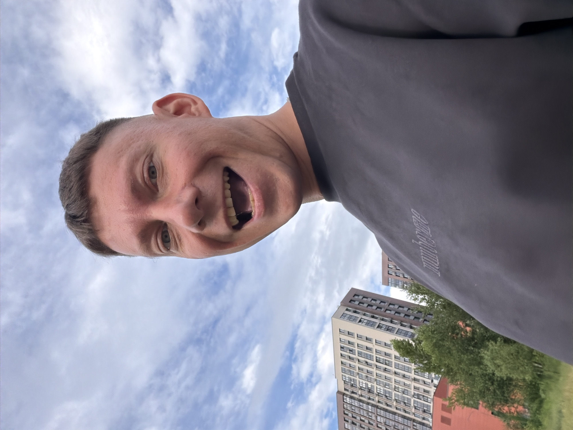 A man with short, buzzed hair and an open-mouthed smile is pictured from the chest up, looking directly at the camera. He is wearing a dark gray sweatshirt with a subtly embroidered logo. The background features a bright, partly cloudy sky, with two tall, modern residential buildings visible on the left. These buildings, characterized by their numerous windows and varying facade colors, are located in a densely populated urban area. The greenery of trees and grass suggests an outdoor setting. The architectural style of the buildings, particularly the multi-story design and window patterns, is consistent with contemporary construction found in Moscow, Russia.