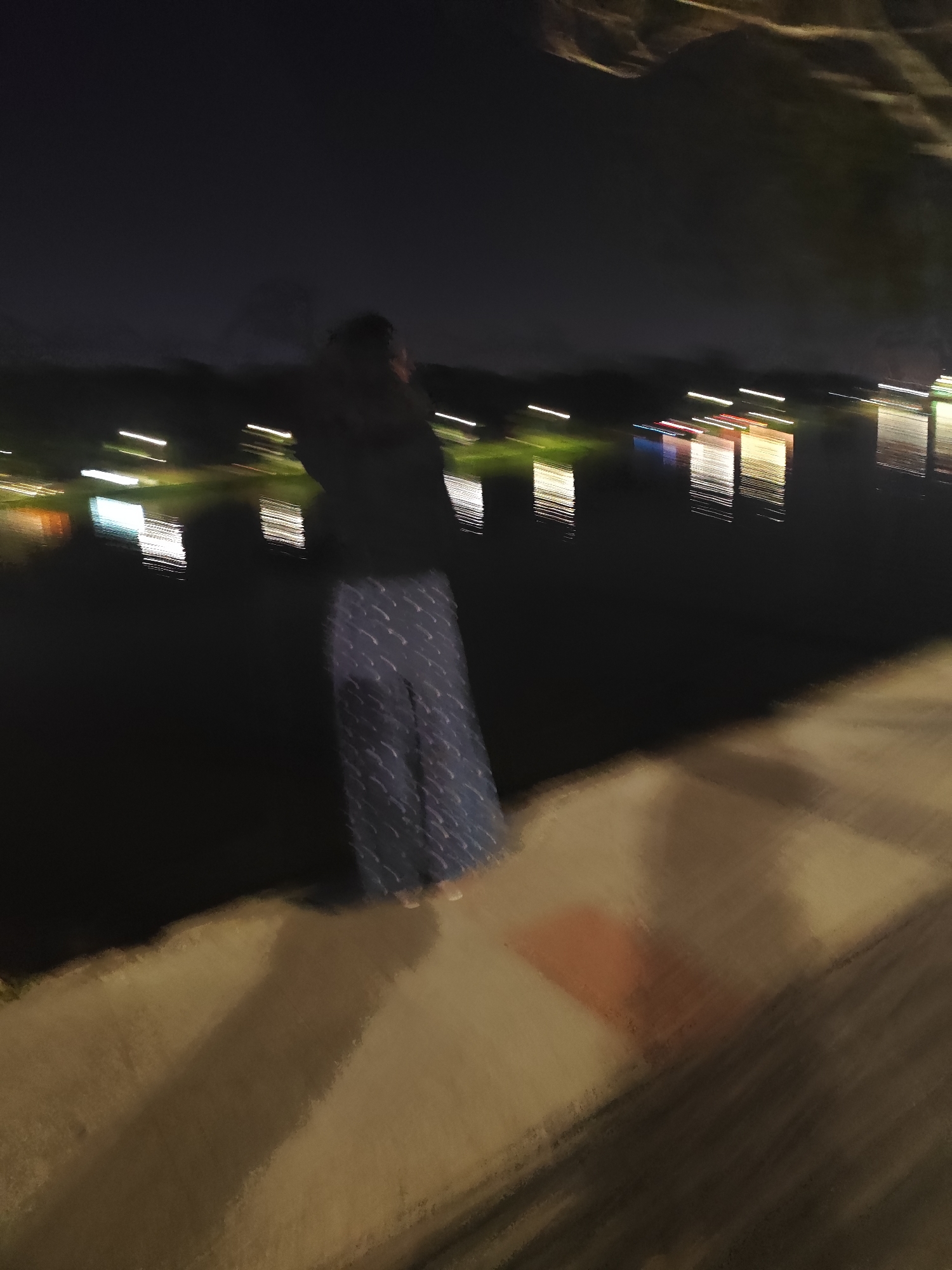 The image shows a blurry, nighttime scene at a body of water, possibly Xuan Huong Lake in Da Lat, Vietnam. A person, seen from behind and partially blurred, is standing on a concrete embankment. The person is wearing dark clothing on their upper body and dark, patterned pants. Lights from buildings or boats are reflected on the water, creating streaks of color. The overall impression is one of motion blur, suggesting either camera movement or movement of the subjects within the scene. The embankment appears to be a pedestrian walkway or viewing area.