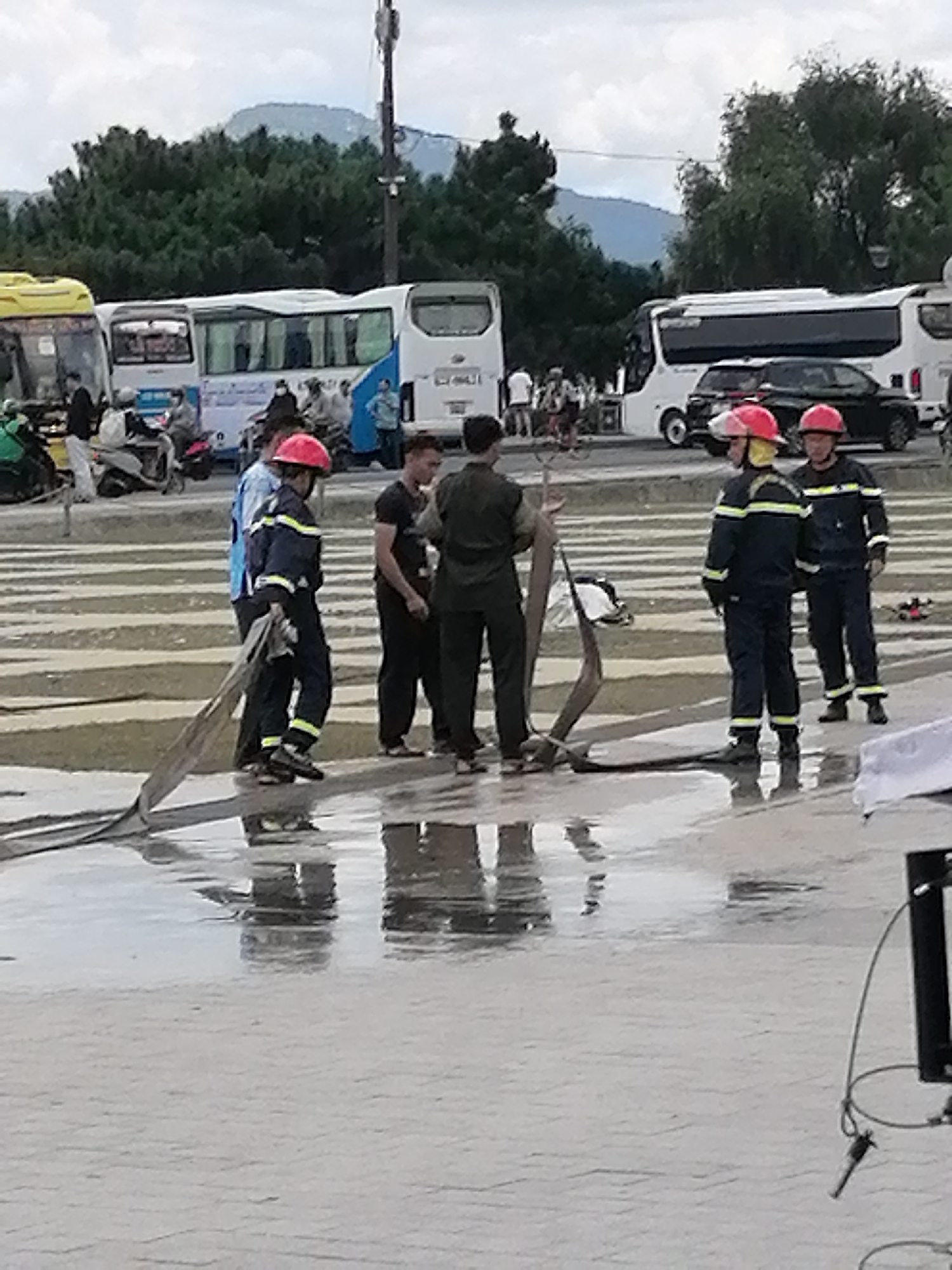 In Da Lat, Vietnam, a group of people, some in what appear to be firefighter uniforms, are gathered around a large hose on a wet, patterned pavement area. The scene takes place on a street or plaza with several large white buses and other vehicles visible in the background. The sky is overcast, and lush green trees and hills are visible in the distance. The foreground is dominated by the wet pavement, reflecting the figures and surrounding objects.