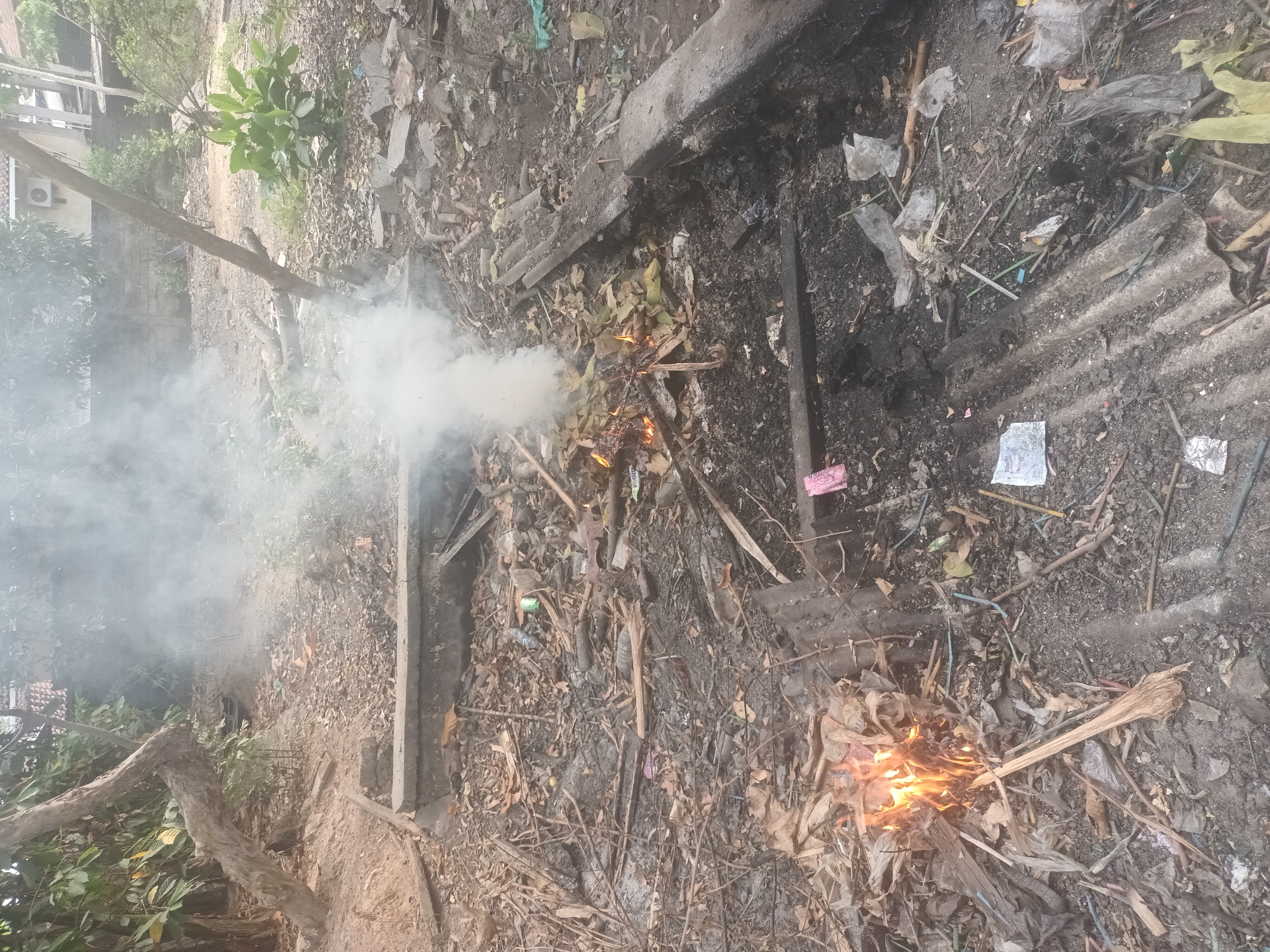 The image depicts an outdoor scene in Mulyo Asri, Indonesia, characterized by an area of discarded debris and a small fire. The ground is covered with a mixture of dry leaves, twigs, and various types of trash, including plastic bottles, bags, and what appears to be corrugated metal sheeting. A fire is actively burning in the lower left portion of the frame, producing visible white smoke that drifts upwards and across the scene. There are no people visible in the image. The surrounding environment includes trees and foliage, suggesting a semi-natural or neglected urban space. The overall impression is one of an unmanaged outdoor area with evidence of burning waste.