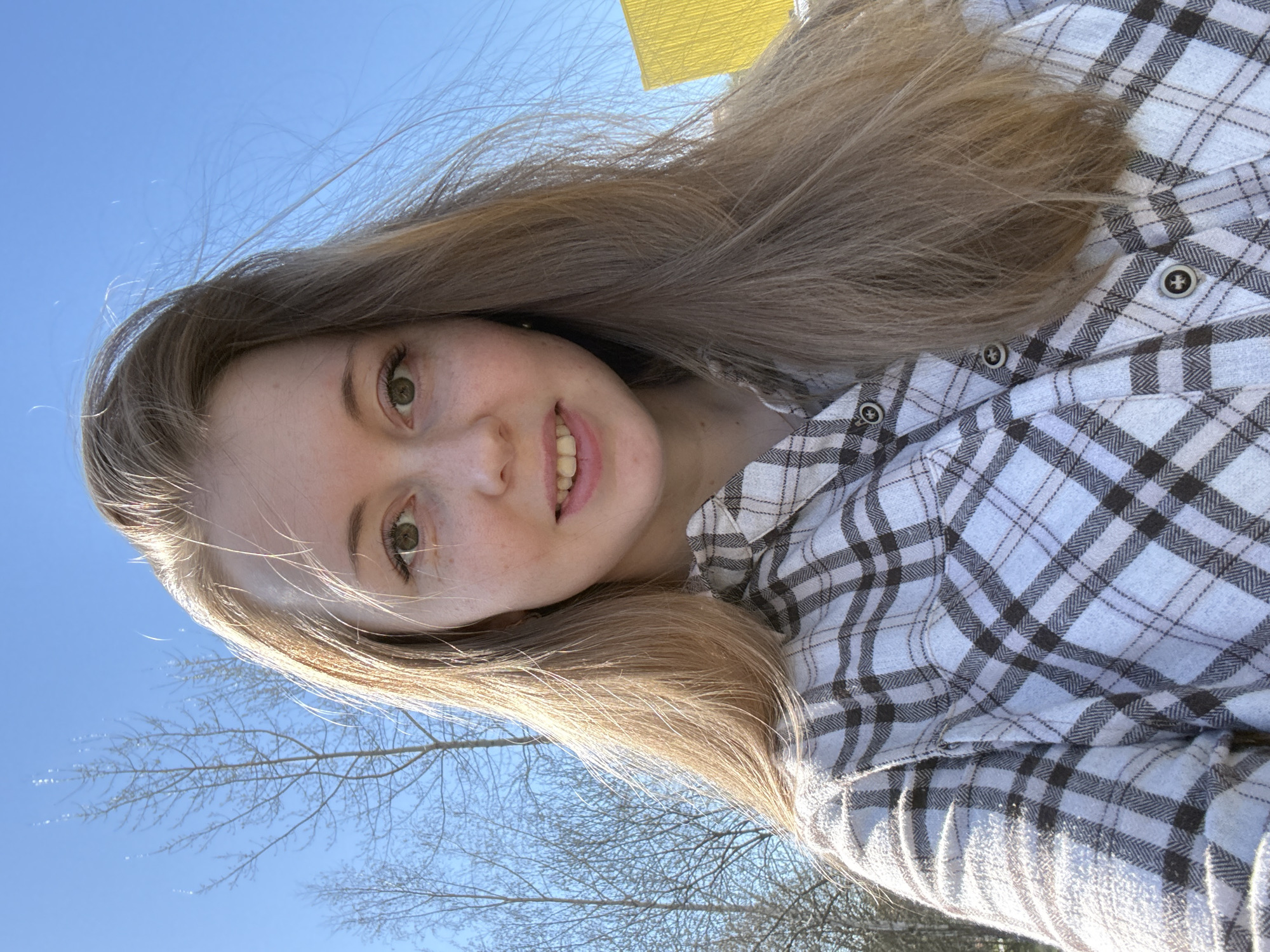 A young woman poses for a selfie outdoors. She has long blonde hair and green eyes. She is wearing a white and black plaid shirt. The background consists of a clear blue sky, a bare tree, and a yellow structure. The exact location in the city of Unknown, Unknown, is not identifiable from the image.