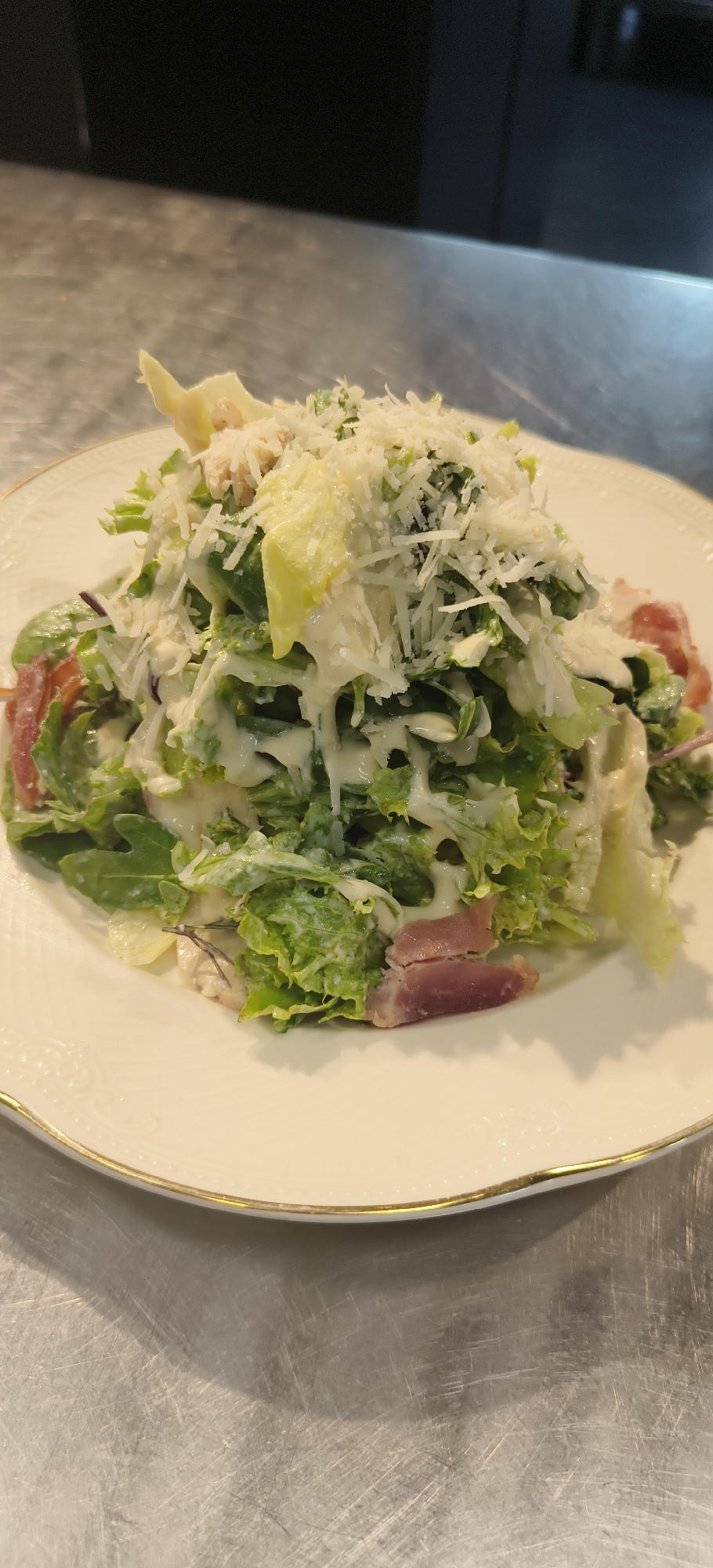 A Caesar salad on a white plate with a gold rim is sitting on a stainless steel surface. The salad is comprised of green lettuce, croutons, parmesan cheese, dressing, and pink meat (likely prosciutto). The plate is centered in the frame. The background is out of focus, showing a black surface above the stainless steel. The image was likely taken indoors under artificial lighting. There is no indication of location in Yerevan, Armenia.
