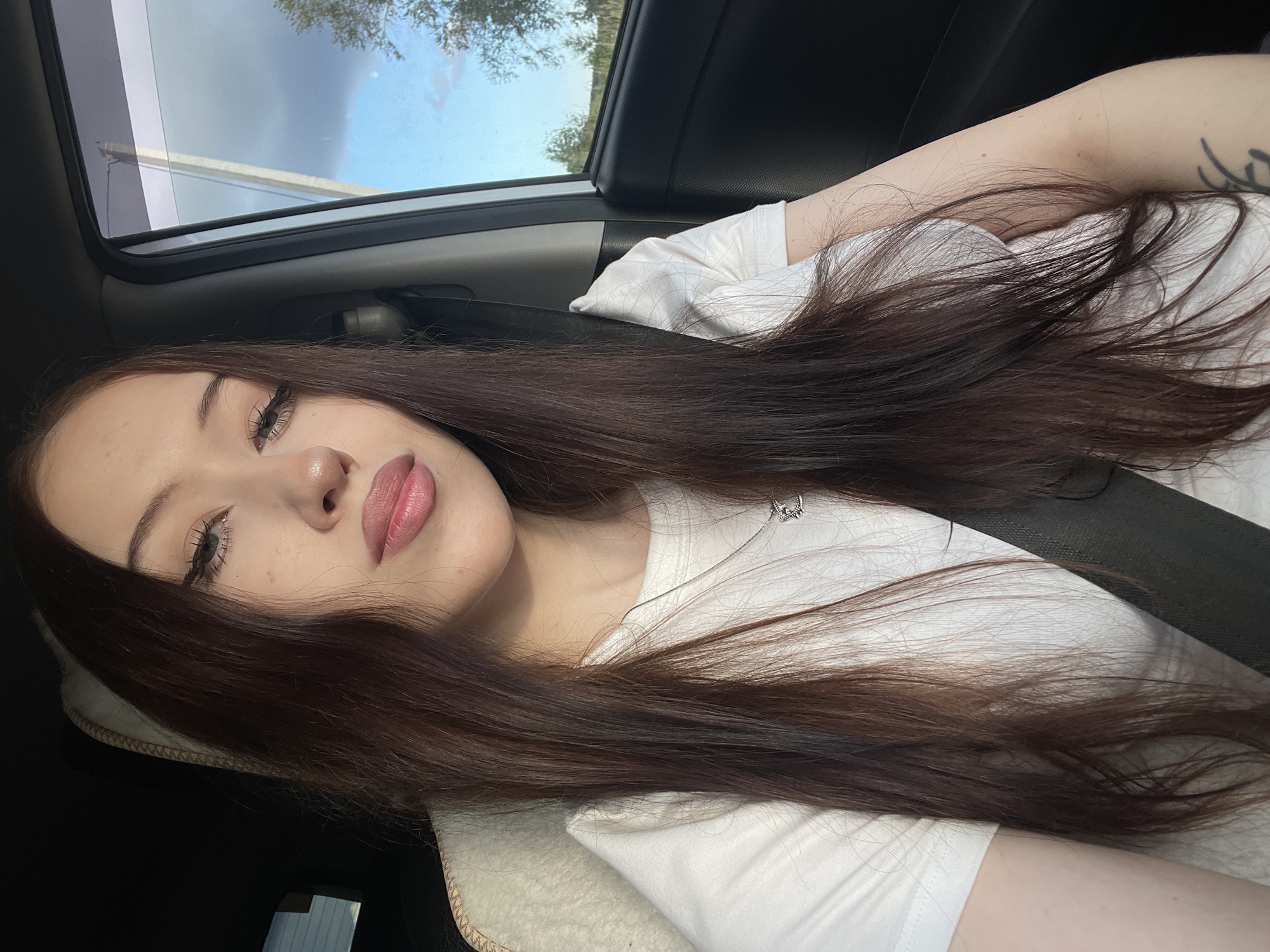 This is a close-up, rotated portrait of a young woman inside a car. She has long, dark brown hair that is spread across her chest and shoulder. Her face is turned towards the camera, with her eyes looking slightly upward. She has noticeable makeup on her eyes, with defined eyelashes and eyeliner, and her lips are painted a soft pink. A delicate silver necklace with a small charm is visible against her white t-shirt. The background shows the interior of a car, with a seatbelt visible. Through the car window and sunroof, a cloudy sky with some trees can be seen, suggesting it is daytime. The overall mood appears relaxed and casual.