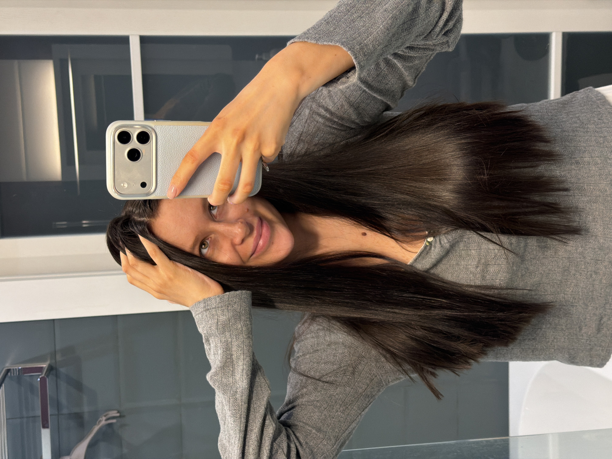 A woman is taking a selfie with a phone that has a gray case. She is wearing a gray, long-sleeved shirt. She has long, dark hair. The background appears to be a bathroom, with a white sink and gray tiled wall. A window with white frames is visible in the background. There is no information about the location being in the city of Khimki, Russia, or any other geographical details.