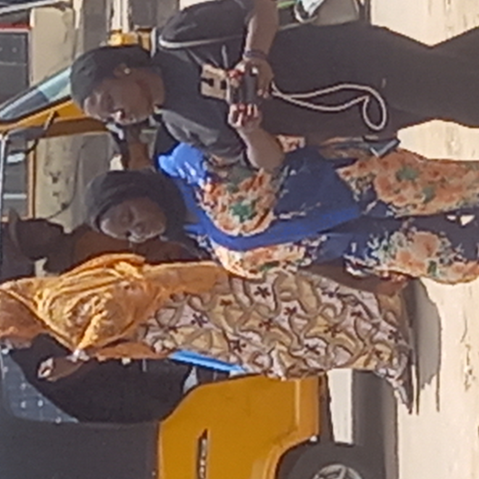 The image captures a daytime scene in Sardauna, Nigeria, featuring several individuals and a yellow three-wheeled auto-rickshaw.
In the center of the frame, within the vehicle, a person is resting, partially covered by a patterned orange and yellow fabric. Adjacent to them, another individual is seated, wearing a blue top with a vibrant floral or abstract pattern in shades of orange, green, and white, complemented by matching patterned pants. This person also wears a dark head covering.
Standing above and slightly behind these two, another person is visible, dressed in a black t-shirt, dark trousers, and a dark cap. They are holding a smartphone with both hands, directing it downwards, possibly capturing a photograph or video. A white cord or strap hangs from their neck, and a dark bracelet is on their wrist.
A fourth person, in dark clothing, is partially visible on the far left, seemingly in the driver's seat of the auto-rickshaw. The vehicle is predominantly yellow, with black tires and some light blue structural elements. The background features light-colored walls or buildings, indicating an outdoor, possibly urban, setting. The scene is brightly lit by natural light, suggesting a sunny day. No discernible text is visible on any subject or object. The overall impression is that of a casual street moment or a pause in travel.