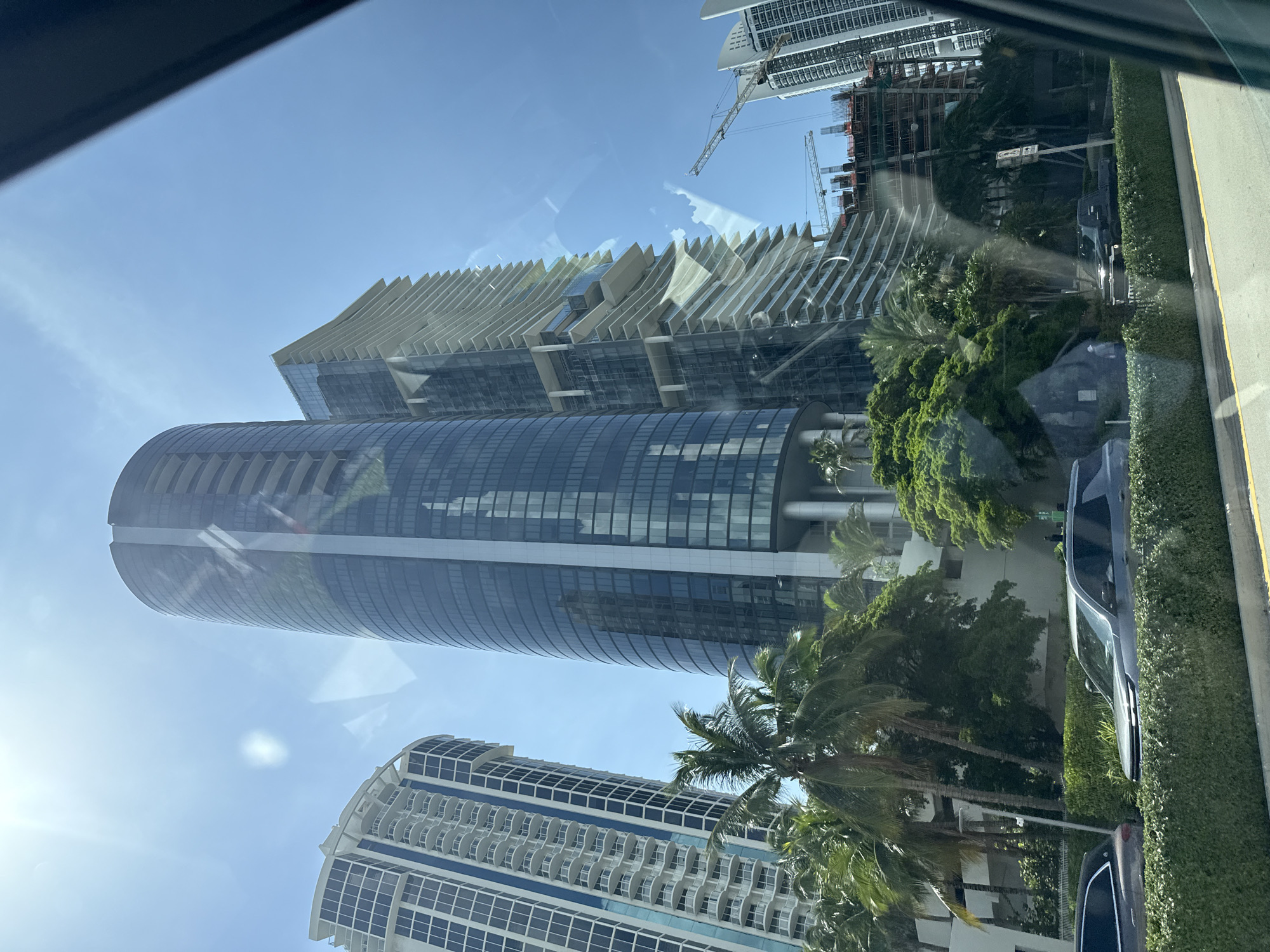 The image shows a street-level view of several modern skyscrapers in Sunny Isles Beach, Florida, USA. In the foreground, a well-maintained green hedge lines the street, with a gray SUV and a darker vehicle parked behind it. Palm trees and lush green foliage are visible between the hedge and the buildings.
Dominating the scene are three prominent skyscrapers. The central and largest is a tall, cylindrical building with a dark, reflective glass facade and vertical white accents. To its left stands a narrower, white skyscraper with a grid of windows and balconies. To the right of the cylindrical building is a strikingly modern skyscraper with a tiered, undulating design featuring numerous balconies and white horizontal elements.
In the background, to the right, a tall building under construction is visible, with scaffolding and cranes indicating ongoing work. Another white skyscraper, similar in design to the one on the left but taller, is seen further in the distance. The sky is a clear blue with some wispy white clouds, and bright sunlight creates reflections on the glass surfaces of the buildings. The image appears to be taken from inside a vehicle, with a dark car window frame visible in the upper right corner.