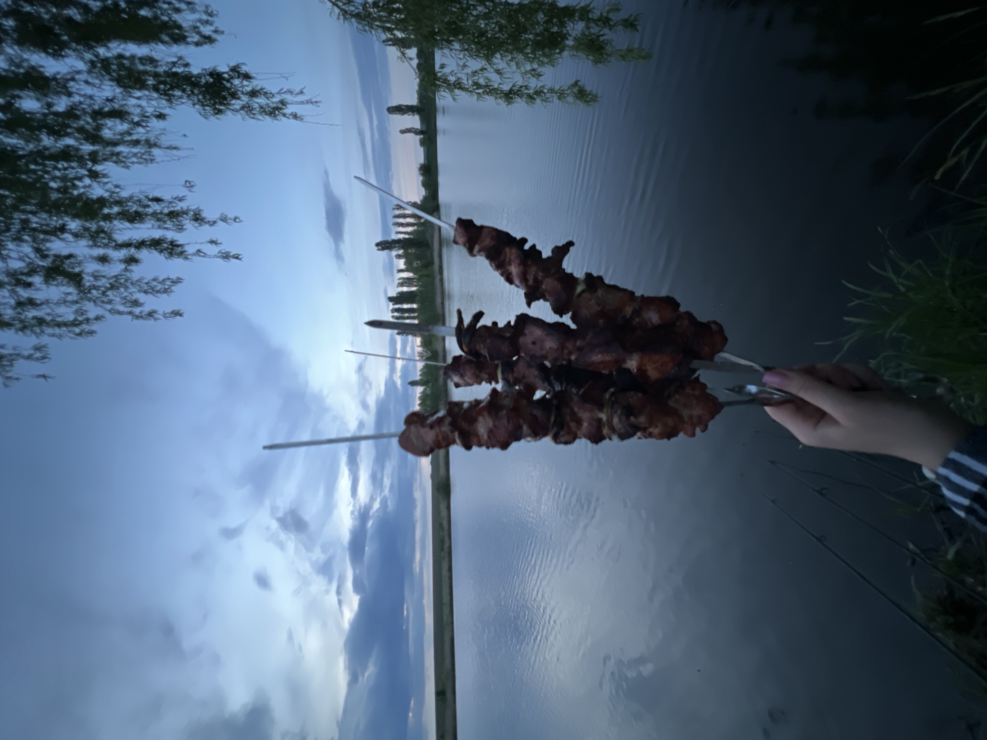 Three metal skewers are held in a person's hand, each laden with cooked meat, likely shish kebab or similar grilled meat. The background depicts a body of water, possibly a lake or wide river, with trees lining the opposite bank. The sky is a mixture of blue and cloud cover, suggesting either dawn or dusk. The location is Romanove Selo, Ukraine. The scene is generally serene, with the focus on the food being presented.