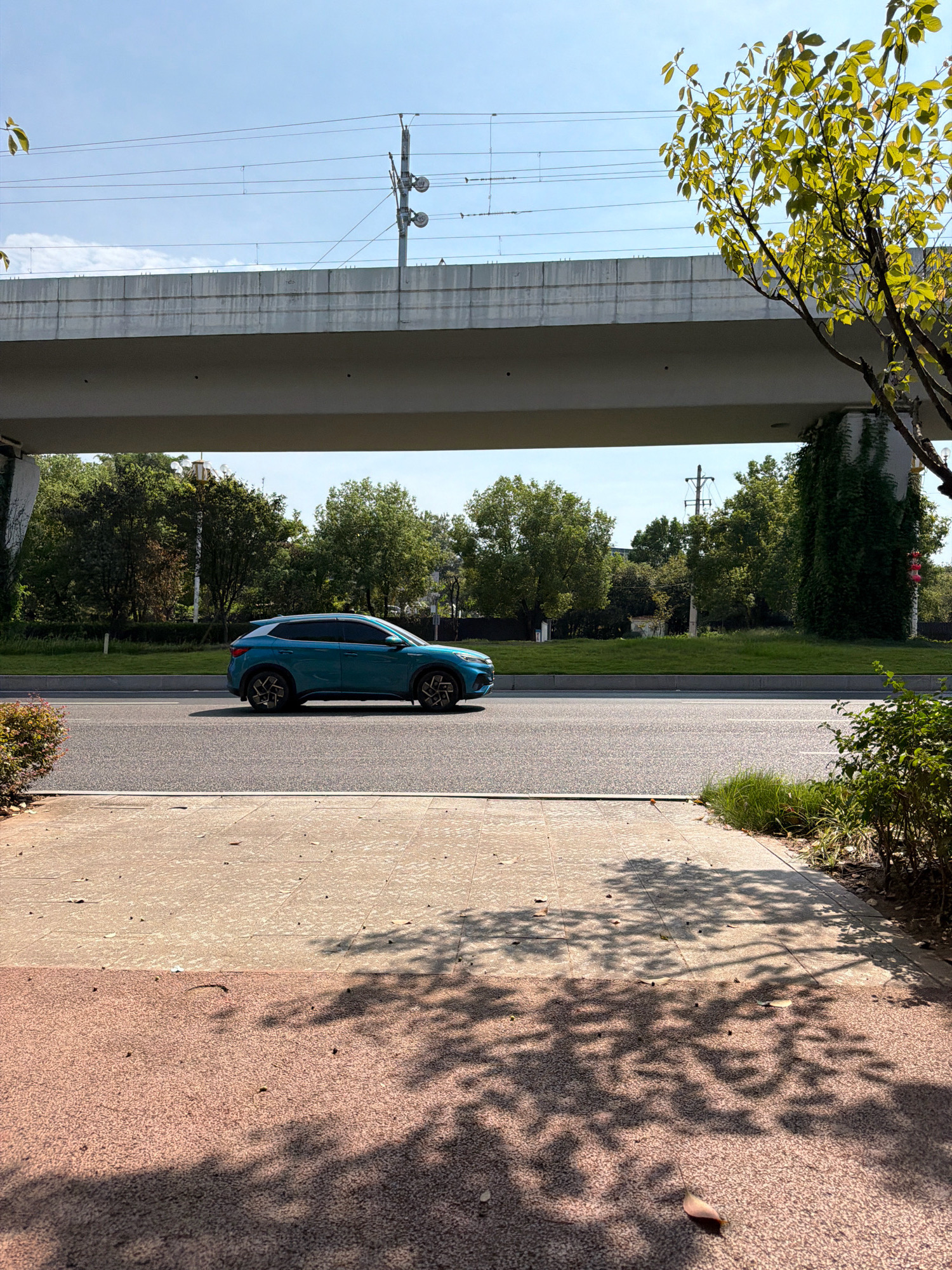 A turquoise SUV drives on a paved road under a concrete overpass. The overpass has overhead power lines running across it. The road is bordered by a grassy median and trees on both sides. In the foreground, a sidewalk made of light-colored paving stones leads to a darker, reddish-brown path, both of which are partially covered in the dappled shadows of tree leaves. The scene appears to be in a city park or a suburban area. This image was taken in Jiangjin District, Chongqing, China.