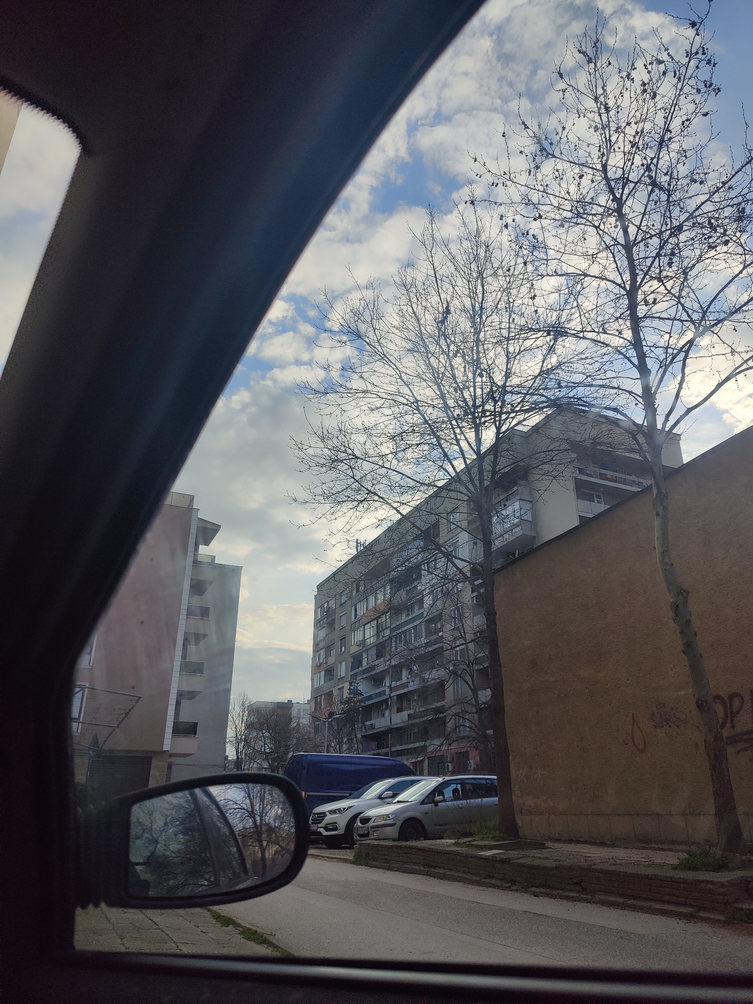 The image displays a static view of a residential street in Plovdiv, Bulgaria, captured from within a vehicle, looking out the passenger-side window. The dark frame of the vehicle's interior, likely the A-pillar and roofline, diagonally bisects the upper left portion of the frame. A convex side mirror on the lower left reflects an indistinct scene of bare trees and the vehicle's interior.
The sky overhead is partially cloudy, showing patches of blue sky interspersed with white cumulus clouds. Multiple bare deciduous trees, consistent with late autumn or winter, are present throughout the scene; two prominent examples stand in the right midground, their branches silhouetted against the sky.
On the left, a modern, multi-story residential building with a light-colored facade features several balconies with white railings and an angular, stepped upper section. In the center-background, a taller, older-style grey multi-story apartment building with numerous windows and balconies is visible. To the immediate right, a low, mustard-yellow colored wall runs parallel to the street, exhibiting faint red graffiti, including the letters "OP".
The paved street in the foreground features a dark blue van parked consecutively behind a silver minivan on the right side. A low, dark-colored concrete or brick barrier separates the street from the base of the yellow wall. No individuals are present or engaged in any actions within the depicted scene.