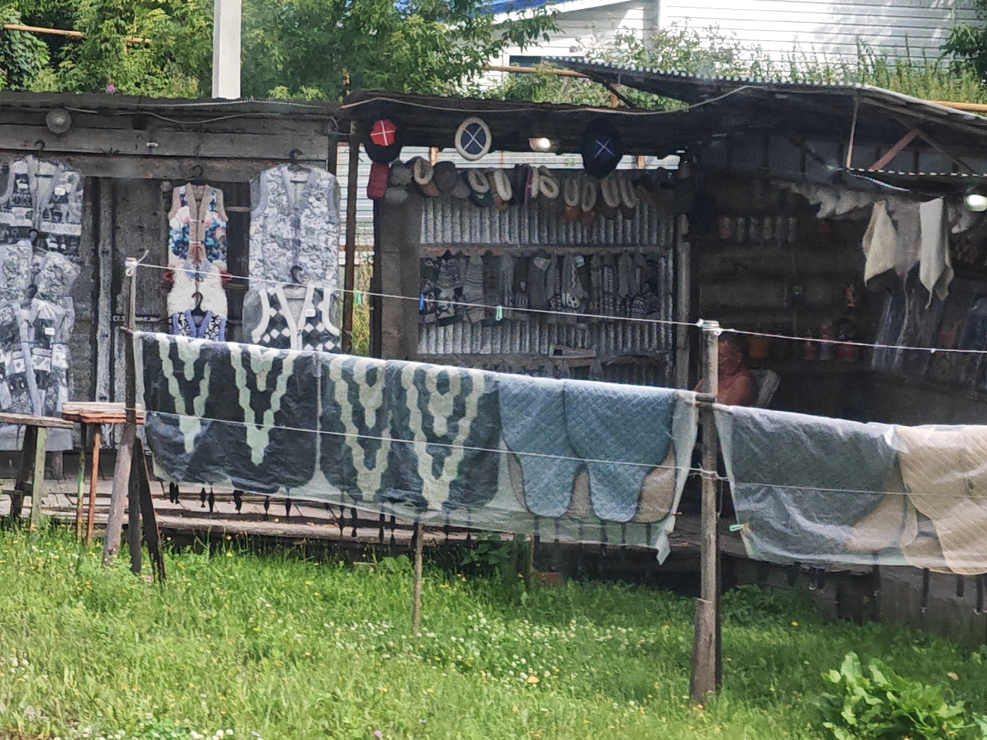 This image depicts an outdoor stall in Novye Vyselki, Russia, featuring various items for sale, including clothing and what appear to be household textiles. The stall is constructed with corrugated metal and wood, with a tarp-like roof providing shelter. On the left side of the stall, several pieces of outerwear, possibly vests or jackets, are displayed on hangers. They have a grey, patterned design. In the center, a colorful vest with floral patterns and traditional fasteners is hung, with additional items below it. To the right, more clothing items and some circular objects, possibly hats or decorative items with symbols, are visible. Shelves behind the stall display rows of what appear to be small containers or bottles. In the foreground, several large textiles with camouflage and blue patterned designs are draped over a railing, suggesting they are also for sale. The ground in front of the stall is covered in green grass with scattered small white and yellow wildflowers. A person, partially visible, is seated behind the stall, with their upper body in view. The overall scene suggests a small roadside market.