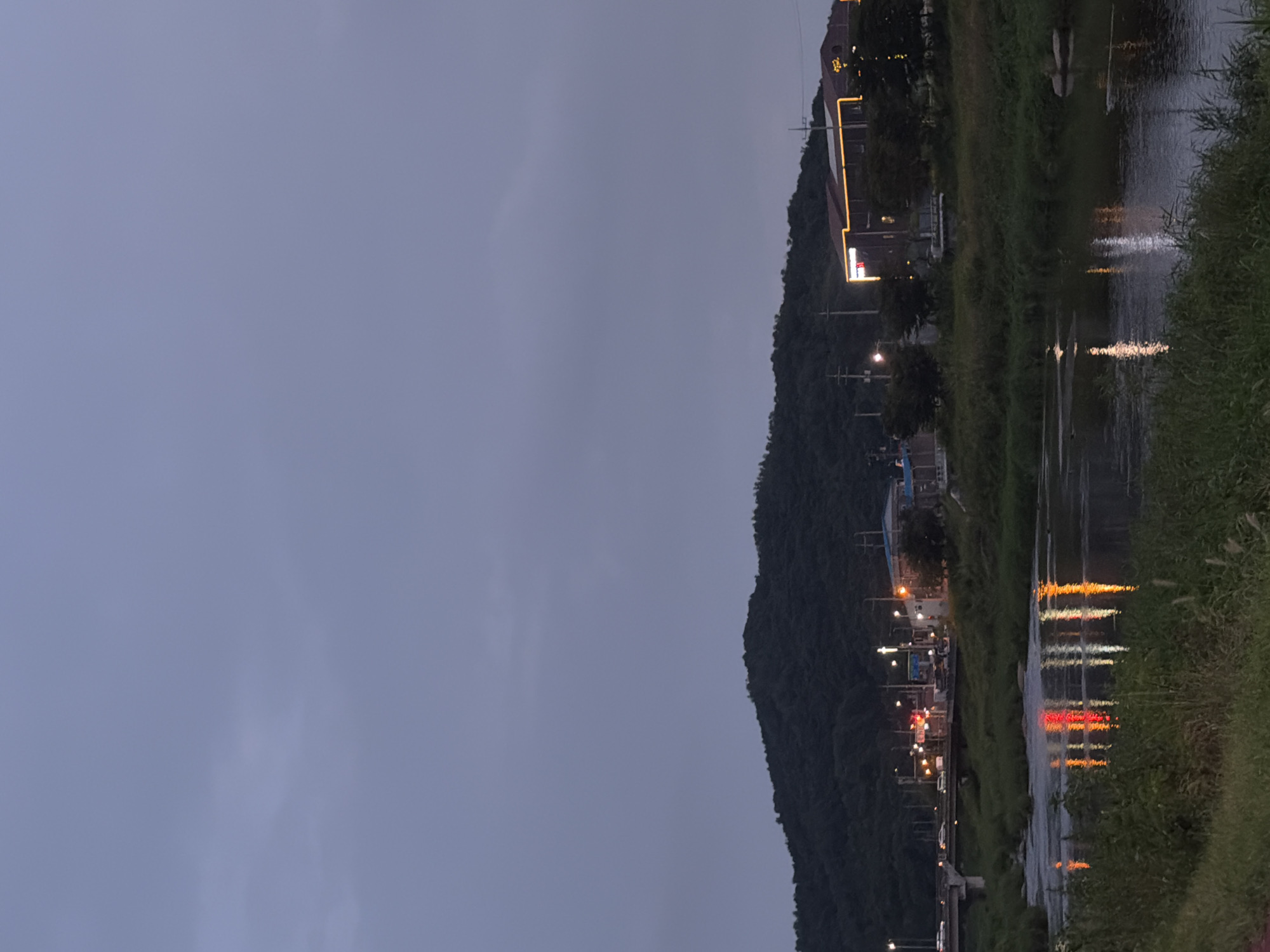 The image captures a dusk scene near a river in Dongducheon-si, South Korea. A dark, tree-covered hill dominates the background. In the middle ground, a bridge crosses the river, with vehicles visible on it. Along the riverbank, there are buildings with illuminated signs and streetlights, reflecting in the calm water. The foreground consists of lush green grass and vegetation bordering the river. The sky is overcast and dark, suggesting the transition from day to night.