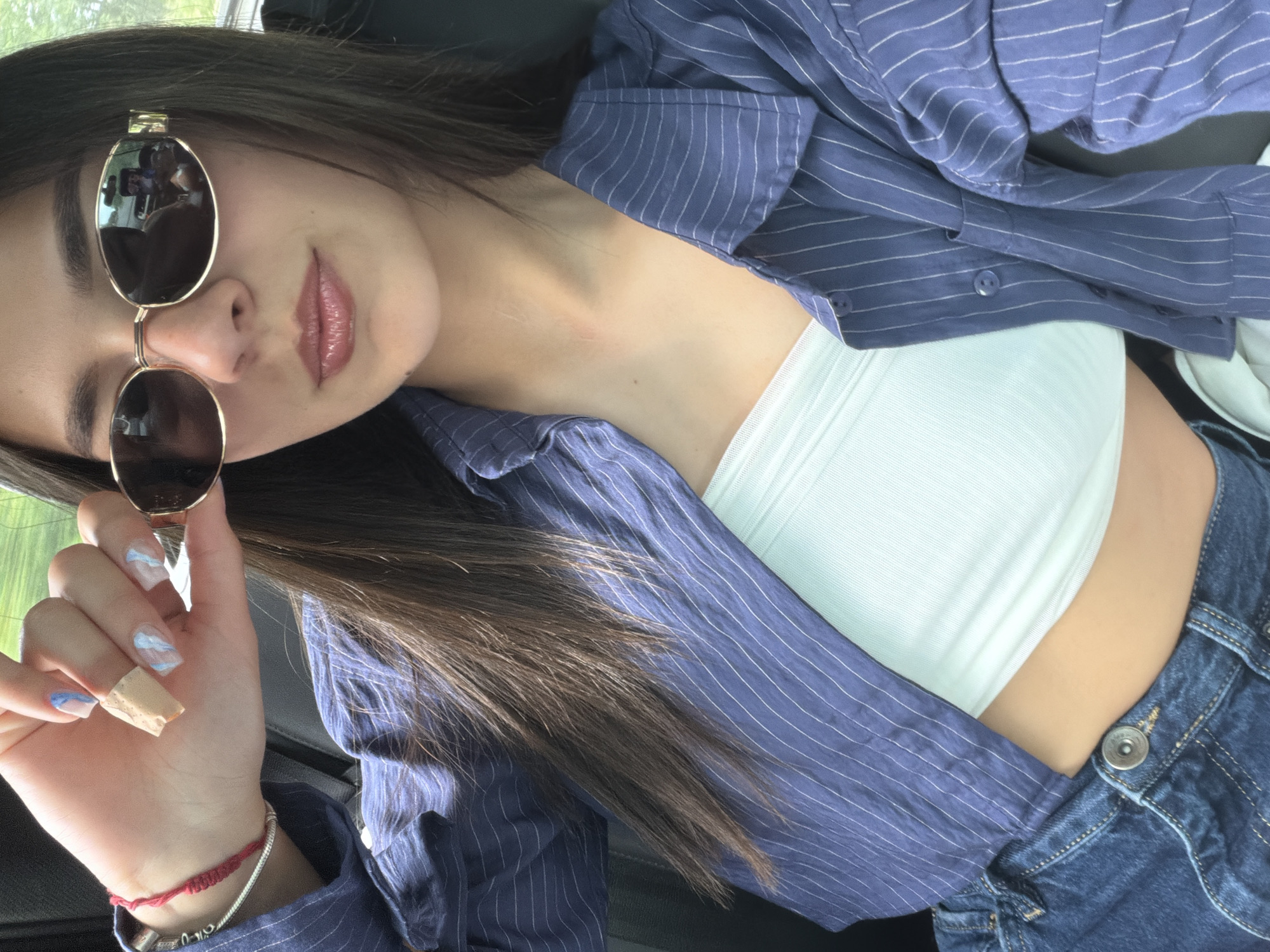 A medium shot depicts a young woman in a seated position, presumably inside a vehicle. She wears dark, oval-shaped sunglasses with a gold frame. The woman's dark hair is parted on the left. Her upper body is attired in a white tube top, overlaid by a blue, pinstriped button-down shirt. The lower portion of her body is clad in denim jeans. The woman's left hand is raised, holding the sunglasses. The visible nails on her left hand are decorated with light blue and white nail polish, except for one finger with a bandage. Two bracelets adorn her left wrist. The background suggests the interior of a car, with dark upholstery visible. The location is not discernible from the image and cannot be inferred as Polski Trambesh, Bulgaria.