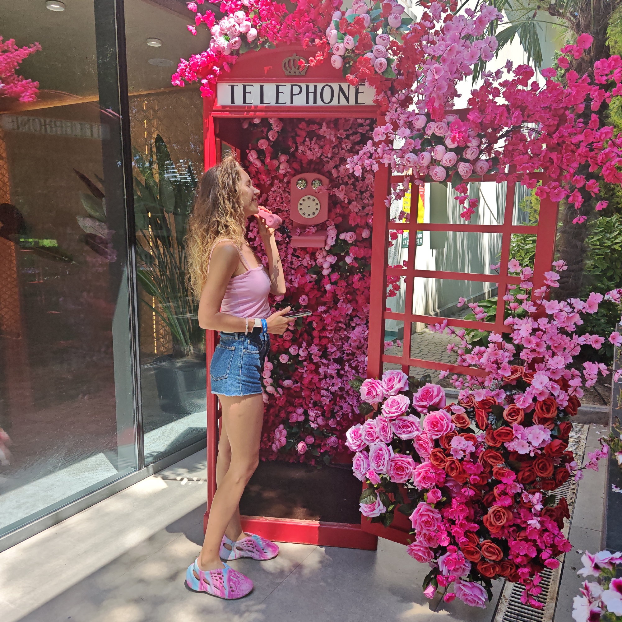 A woman with curly blonde hair stands to the left of a red telephone booth that is decorated with abundant pink and red flowers. The woman wears a pink tank top, blue denim shorts, and pink and blue patterned slip-on shoes. She holds a mobile phone in her right hand and a pink phone receiver to her ear with her left hand. She is looking to her left, up and away from the camera. The telephone booth is covered in faux flowers, and a pink rotary phone is mounted on the inside back wall. The telephone booth is positioned in an outdoor setting, with a glass building with reflections to the left and foliage and a brick sidewalk to the right. Sunlight illuminates the scene.