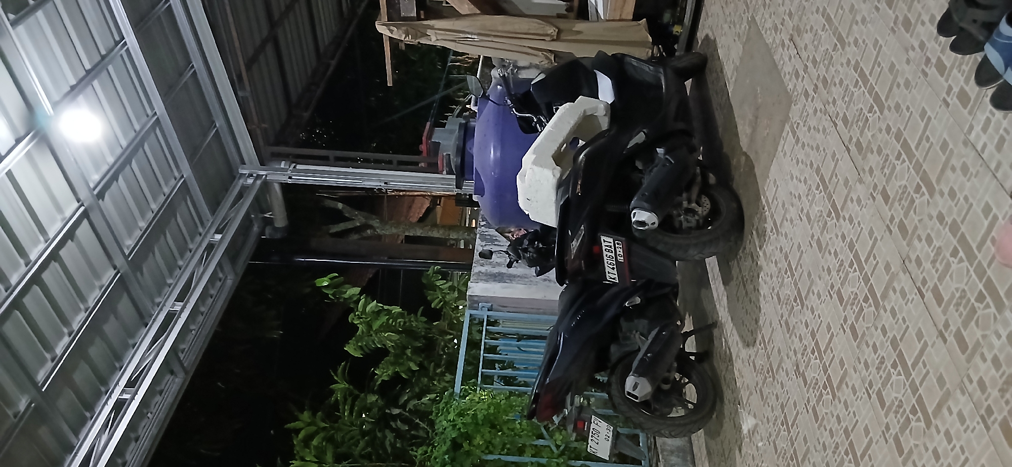 Two dark-colored motorcycles are parked side-by-side under a metal awning, likely in a residential area of Samarinda, Indonesia. The motorcycle in the foreground has the license plate "KT 4616 BAT" and "10-28." The motorcycle in the background has the license plate "KT 2750 FV" and "02-30." A large, purple water tank is partially visible behind the motorcycles, along with a beige cloth hanging on a wooden structure. Green foliage is visible on the left side of the image, and a blue metal fence is present. The ground is paved with patterned tiles. A bright light illuminates the scene from the metal awning.