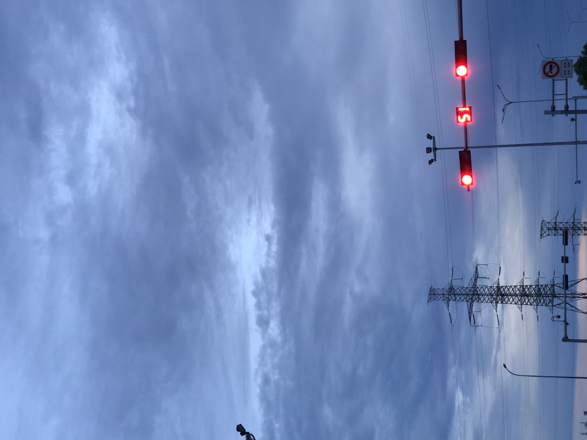 This media file shows a traffic light with a digital timer displaying "15" and two red lights illuminated, indicating that traffic is stopped. The traffic light is positioned on a pole with several wires extending from it, suggesting an urban or semi-urban setting. In the background, a large, intricate power transmission tower is visible, with numerous high-voltage lines stretching across the frame. The sky is overcast with dramatic, textured clouds, hinting at a potentially stormy or twilight atmosphere. To the right of the traffic light, a sign with a red circle and a diagonal line through a horn symbol is mounted on a pole, which likely signifies a "no honking" rule. The overall scene appears to be a street or roadside in Đà Nẵng, Vietnam, at dusk or during a period of inclement weather. There are no people visible in the frame.