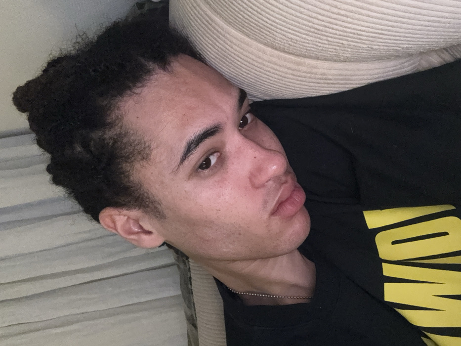 A light-skinned person with dark, curly hair lies against a striped surface, likely a couch or bed. The person wears a black shirt with a yellow design and a silver chain. Their head rests against a beige pillow. The person's expression features slightly pursed lips, appearing thoughtful or contemplative. Light illuminates their face, highlighting subtle freckles and features.