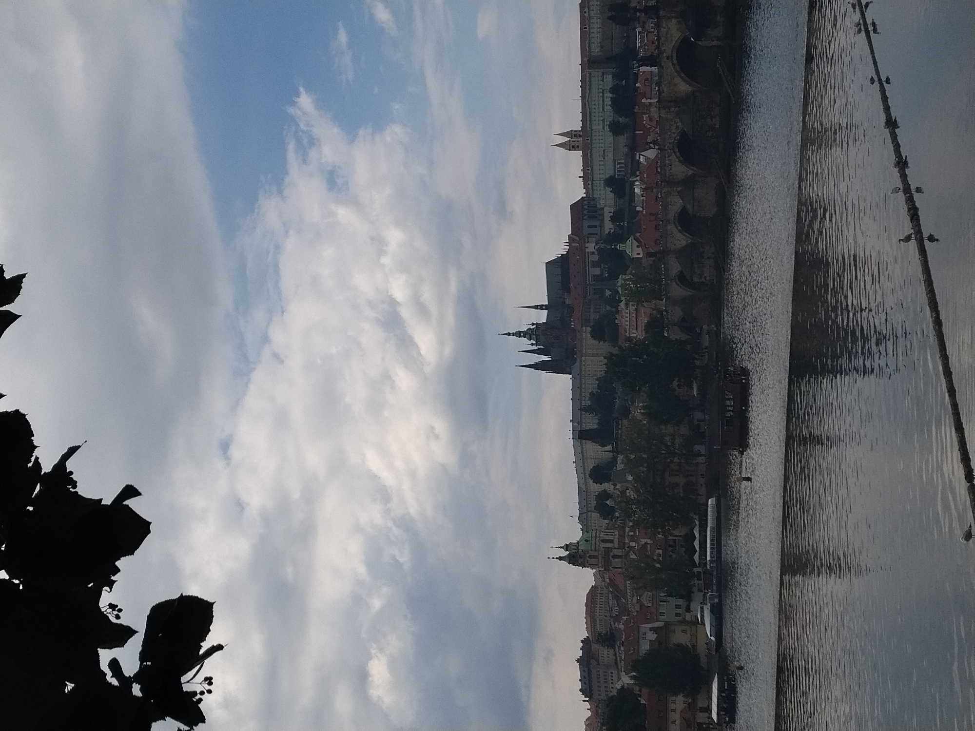The image depicts a panoramic view of Prague, Czechia, with Prague Castle and St. Vitus Cathedral prominently situated on a hill overlooking the Vltava River. The sky is overcast with dramatic clouds, interspersed with patches of blue. In the foreground, the Vltava River flows, with its surface reflecting the sky and the cityscape. Several boats are visible on the river, including a tour boat and some smaller vessels. A line of arched stone bridges spans the river, with Charles Bridge being the most recognizable. The architecture of Prague is evident, with numerous red-tiled roofs and spires characteristic of the city. The overall scene conveys a sense of historical grandeur and a slightly moody atmosphere due to the cloudy sky.