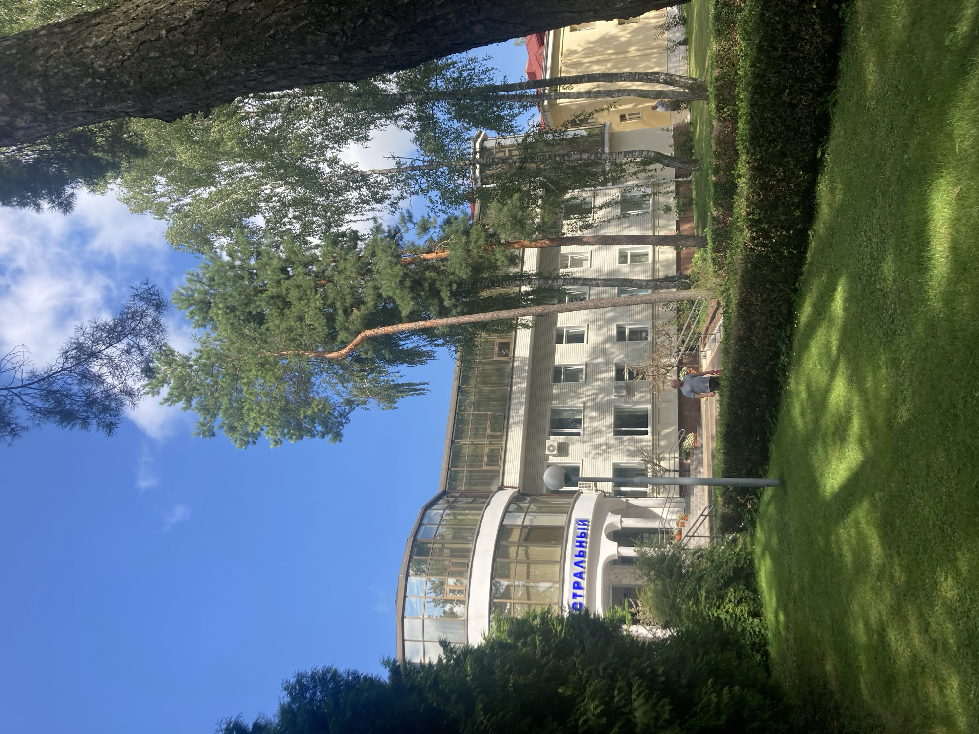 The image shows a building with a curved, glass-enclosed upper floor, with the word "ЕСТРАЛЬНЫЙ" (ESTRALNY) displayed in blue letters above the entrance. The building is light-colored with multiple windows. In front of the building, there is a well-maintained lawn and a trimmed hedge. Several tall trees, including pine and birch, are visible, casting shadows on the grass. A person is walking on a path leading away from the building towards the right side of the frame. The sky is blue with scattered clouds. The scene appears to be set in a park-like environment, possibly a sanatorium or health resort, given the presence of the word "ESTRALNY," which can translate to "central" or "insurance" in Russian, and the overall tranquil setting. This building is the "Baranavichy Central City Hospital" in Baranavichy, Belarus.