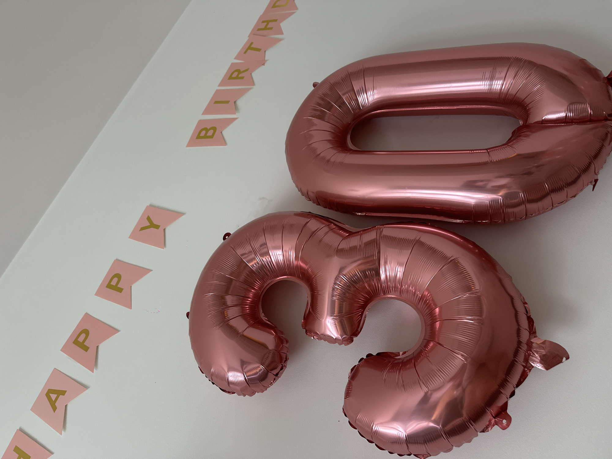A rose gold "3" balloon and a rose gold "0" balloon are positioned on a white wall. Above the balloons, a pink banner with gold lettering spells out "HAPPY BIRTHDAY" in two separate sections. The banner is triangular in shape with each letter on its own flag. The scene is set in a room.