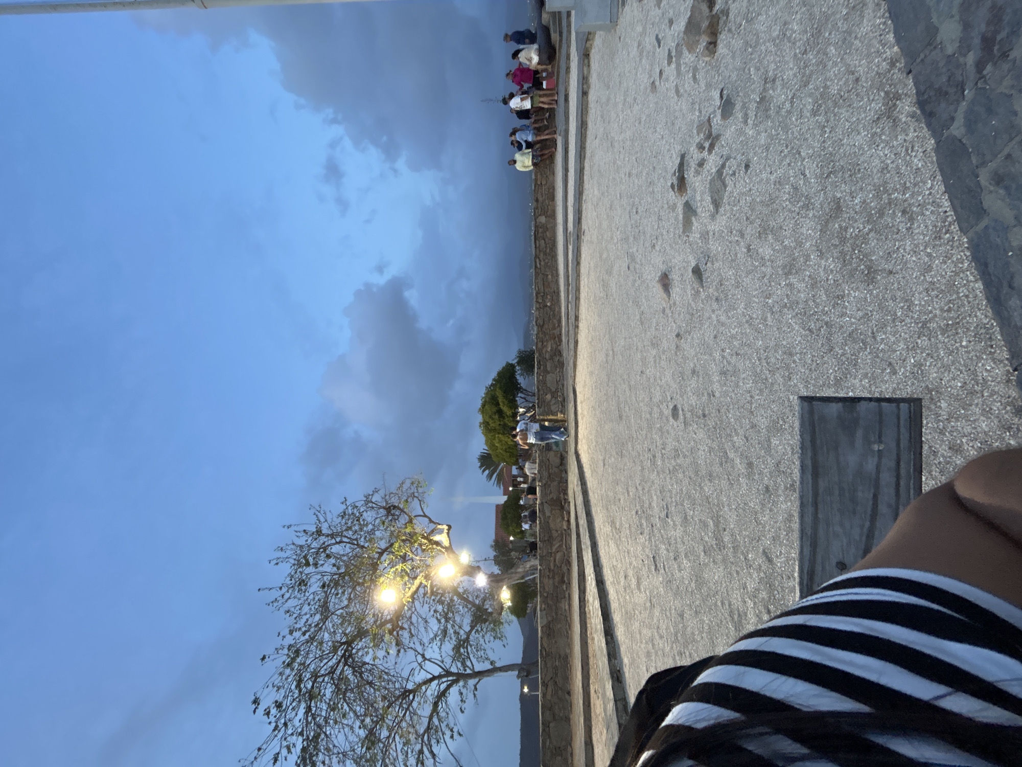 The image captures a twilight scene at what appears to be Fortín La Galera in Juan Griego, Venezuela. In the immediate foreground, the left side shows the torso and arm of a person wearing a black and white striped shirt, seated on a wooden bench. The ground directly in front of the viewer is a light-colored, pebbly surface, transitioning into a larger, open area of similar material. A low, grey stone wall runs horizontally across the midground.
On the left side of the midground, behind the stone wall, two trees are visible. One tree, centrally located, is extensively decorated with yellow string lights, casting a bright glow. Behind the wall to the right of these trees, two individuals are seen walking away from the viewer. Further to the right, a group of approximately ten people are gathered along the stone wall, standing and sitting, observing the panoramic view. A dark, historic cannon is mounted on a stone base at the far right, pointed outwards over the wall.
The background reveals a hazy landscape of hills and mountains, possibly overlooking the Caribbean Sea or a bay, under a vast, deep blue sky. The sky is heavily laden with dark, prominent clouds, particularly on the right side of the frame, indicating the transition from dusk to night. Artificial lighting from the tree lights and distant urban sources contrasts with the dim natural light.