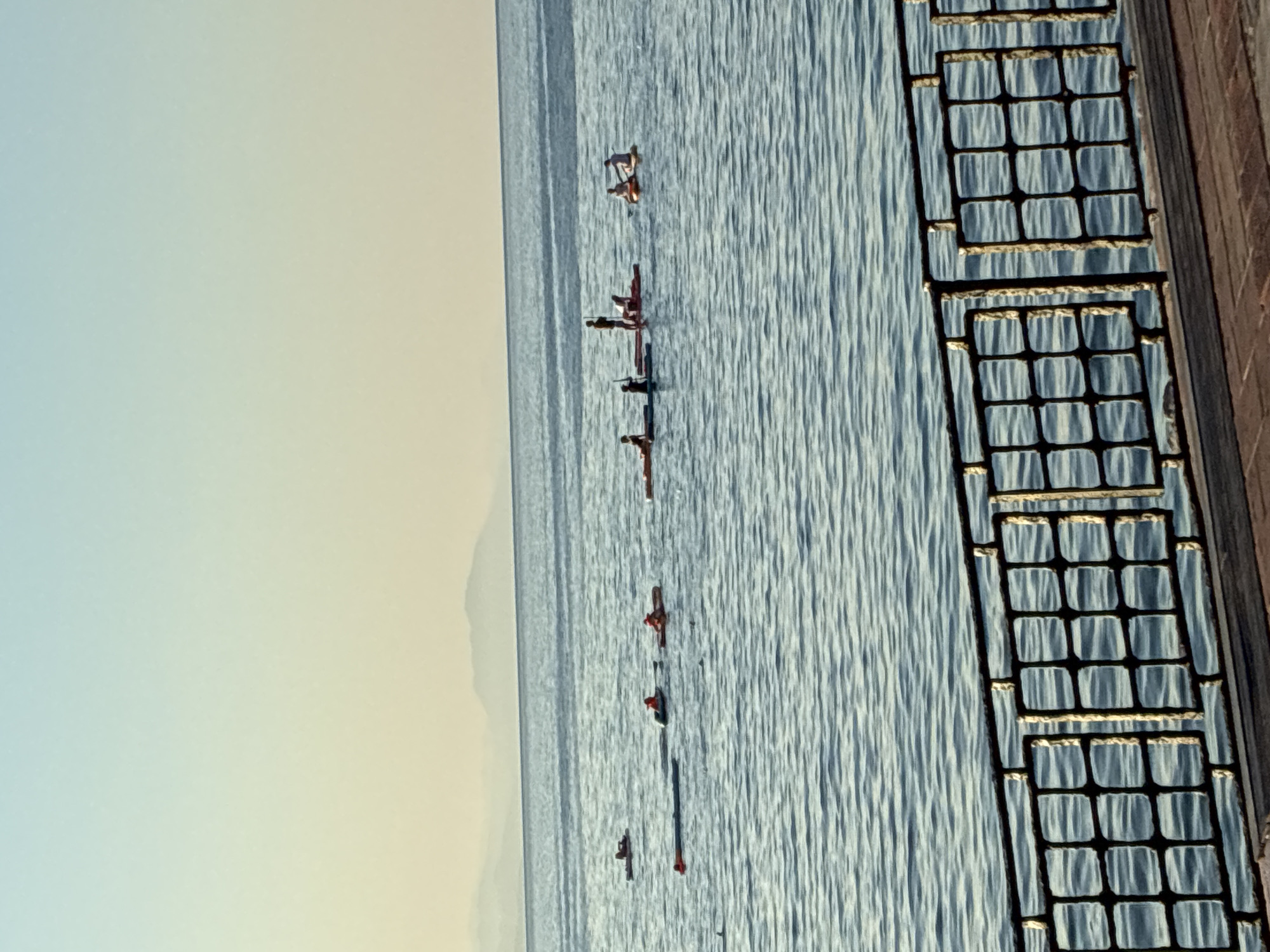 The image shows a wide shot of the sea in Alanya, Turkey, with a clear sky transitioning from light blue at the top to a pale yellow near the horizon. In the distance, hazy mountain ranges are visible. In the foreground, there is a dark metal railing with a repeating square grid pattern, casting a shadow on a wooden boardwalk. The sea is calm with gentle ripples, and several small boats or watercraft are scattered across the water. On these watercraft, multiple individuals can be seen, some paddling or standing. The overall scene depicts a serene coastal environment in Alanya during what appears to be daytime, possibly early morning or late afternoon given the soft lighting.