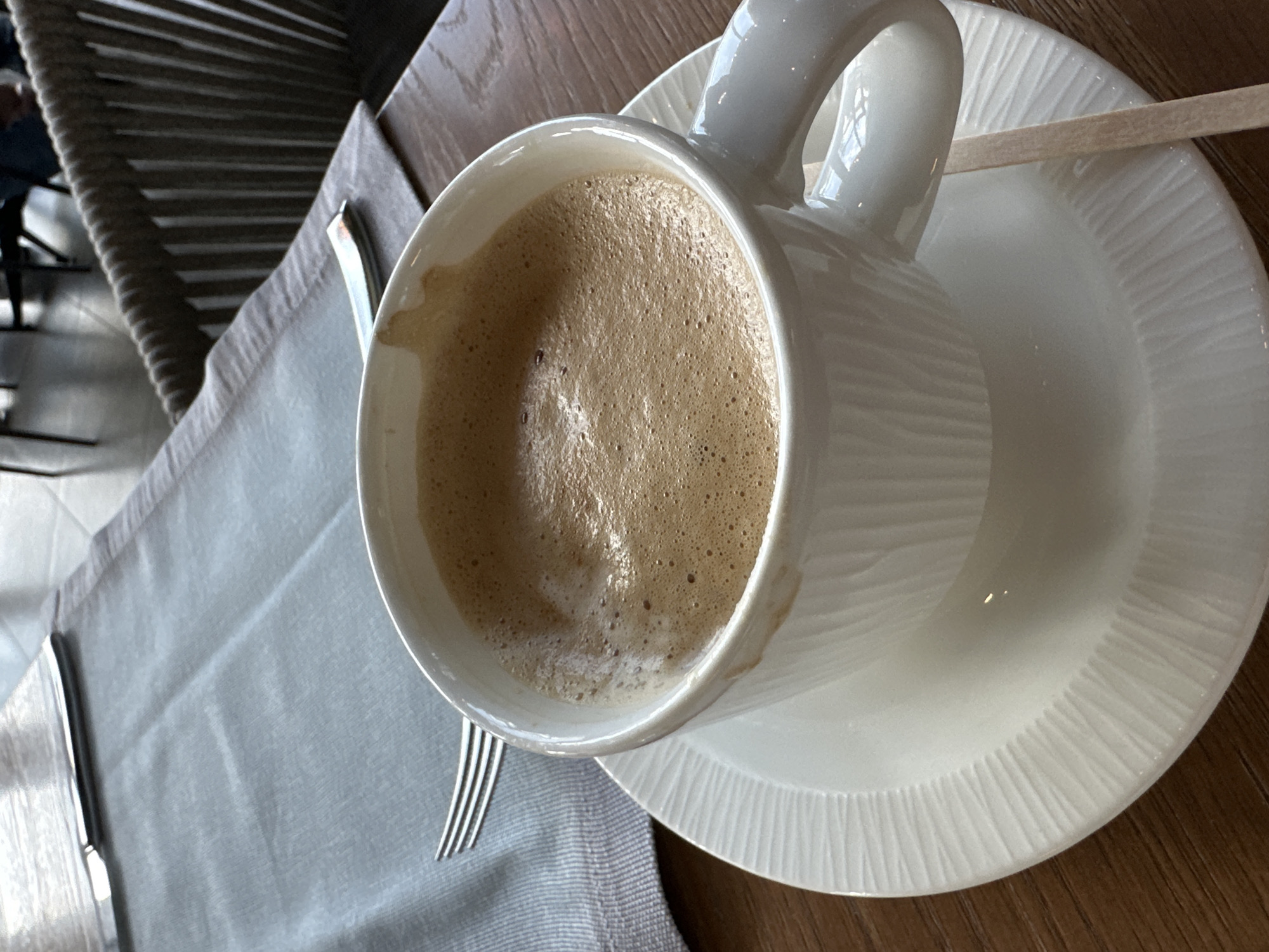The image captures a close-up, overhead view of a white coffee cup filled with a frothy, light brown beverage. The cup rests on a matching white saucer with a textured, ridged pattern. A wooden stirrer lies across the saucer. A table setting is suggested by a gray placemat underneath, partially visible with a fork and knife positioned on it. A chair with a woven back is seen in the background, and the table appears to be made of wood. The specific location in the city of Kazanlak, Bulgaria is not discernible from the image.