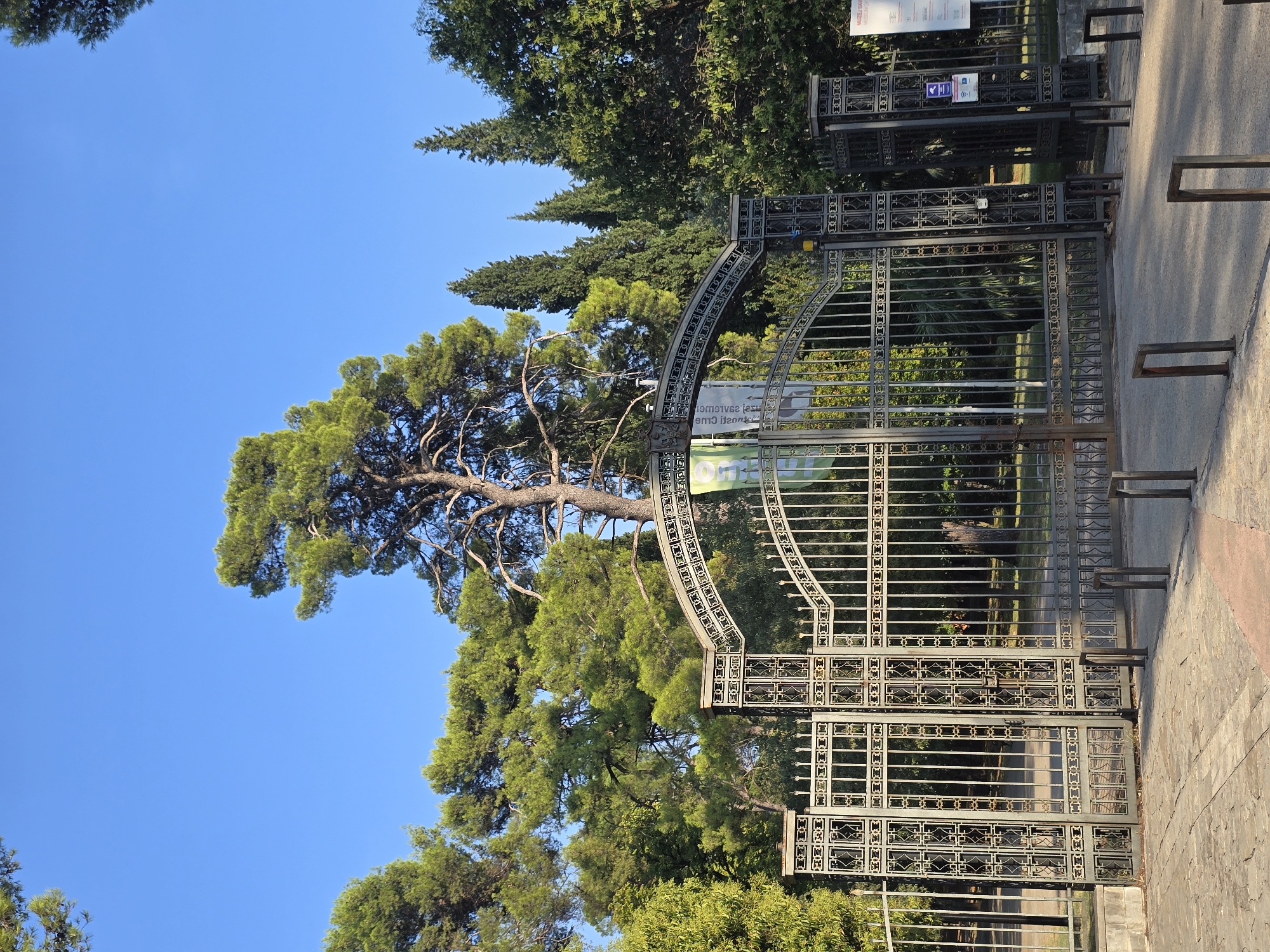 An ornate, tall, wrought-iron gate stands open, admitting passage into a park or botanical garden. The gate features intricate decorative metalwork, with repeating patterns of squares and lines. It is flanked by two substantial, square pillars topped with decorative caps. Behind the gate, lush green trees of various species, including tall pines and cypress-like trees, form a dense backdrop under a clear, bright blue sky. Sunlight casts shadows on the paved ground in front of the gate, suggesting it is morning or late afternoon. A sign is visible hanging from the arch of the gate, and another sign is attached to the pillar on the right side of the gate. The scene appears to be the entrance to the Botanical Garden in Podgorica, Montenegro.