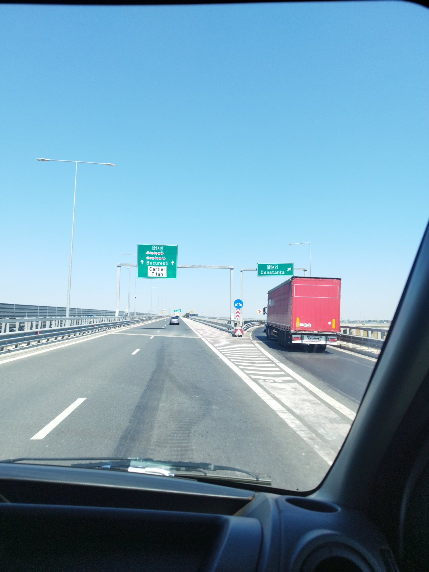 The image depicts a view from inside a vehicle driving on a highway in Glina, Romania, likely on the A2 motorway heading towards Constanța. The sky is clear blue, and the sun is shining brightly. The highway is multi-lane with clear white markings and barriers on the sides. In the foreground, the dashboard and windshield of the vehicle are visible, indicating the driver's perspective.
A large red semi-trailer truck is prominently in the right lane, with the brand "SCHMITZ" visible on its rear. A green highway sign indicates "A2 Constanța" with an arrow pointing forward, suggesting the truck is following the route to Constanța. Further ahead on the left side, another green highway sign displays "A0" followed by names such as "Ploiești", "Urziceni", and "București" with directional arrows. Below these are "Cartier" and "Titan".
The environment appears to be a modern highway infrastructure with streetlights and traffic signs. The road surface shows some tire marks, typical of heavy traffic. In the distance, other vehicles can be seen on the highway. The overall scene conveys a sense of travel and transit on a sunny day.