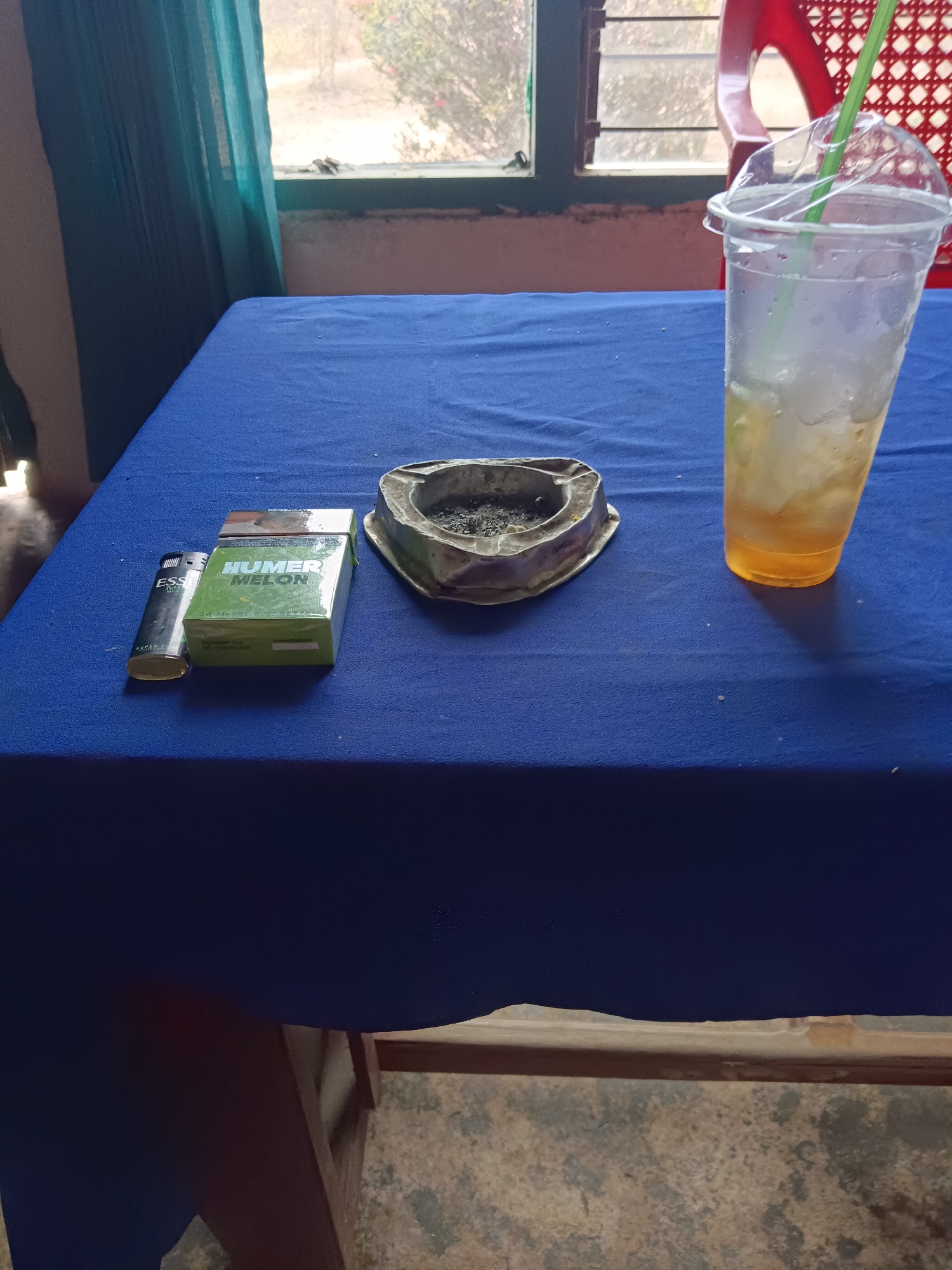 In a room with a blue tablecloth-covered table, a cigarette pack of "HUMER MELON" brand is displayed next to a lighter. An ashtray filled with cigarette butts sits nearby. A plastic cup containing a beverage with ice and a green straw is also present on the table. The scene appears to be located in Tegal Gondo, Indonesia, as indicated by the presence of local cigarette brands and environmental cues. The background shows a window with a view of vegetation and a red chair.