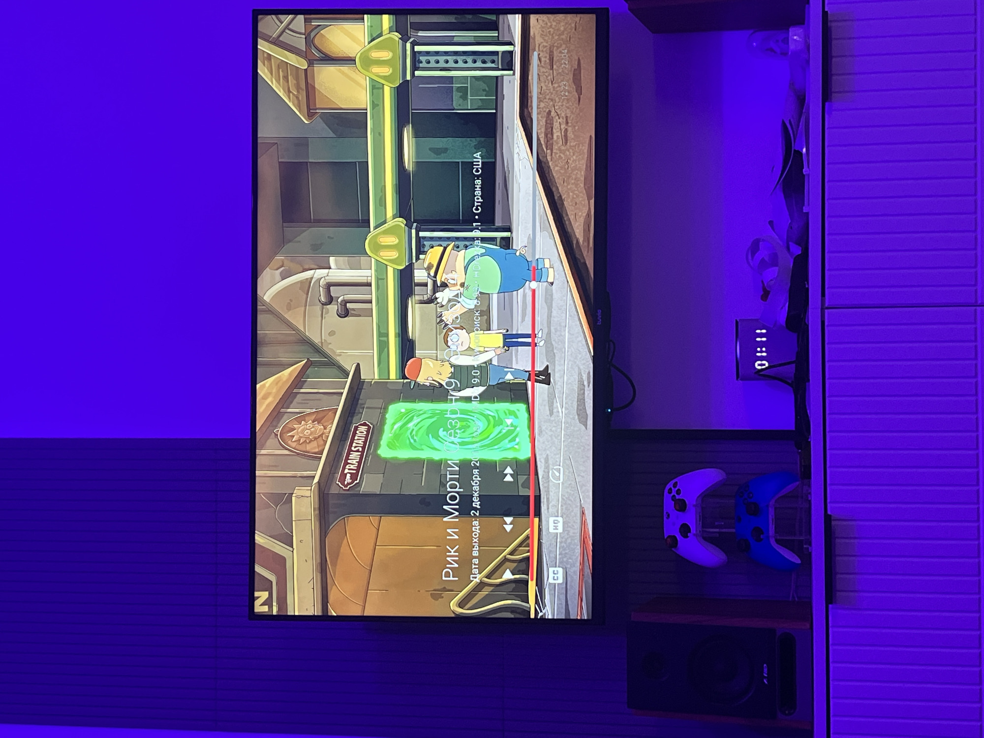 This media file shows a television screen displaying a scene from the animated show "Rick and Morty". The scene depicts the characters Rick and Morty standing in front of a glowing green portal. The setting appears to be a train station, with a sign that reads "TRAIN STATION". There are also details on the screen indicating "Rick and Morty Season 9", "Release Date: December 2, 20", and "Country: USA". The time on a digital clock next to the television shows 01:11. Two gaming controllers, one white and one blue, are mounted on the wall to the right of the television. A speaker is placed below the controllers. The overall lighting in the room is purple, suggesting ambient mood lighting.