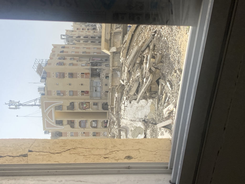 The image, captured from an interior window, depicts a landscape of extensive structural damage in the city of Khan Yunis, Palestinian Territory. The immediate foreground shows the bottom segment of a light-colored window frame, with a vertical crack visible on the left side of the window's interior casing. Beyond the window, the mid-ground is dominated by a large, irregular expanse of rubble, consisting of broken concrete slabs, masonry, and other building debris, indicating severe destruction. In the background, a multi-story residential building with a light tan facade and decorative arched elements is prominently visible. This structure displays significant damage, including numerous broken or missing windows, partially collapsed wall sections, and exposed interiors; some balconies still show hanging laundry. A cellular tower with multiple antennae, along with water tanks and other equipment, is present on the building's rooftop. Further to the right and in the distance, additional multi-story buildings, also appearing damaged, extend into a hazy, bright sky. No individuals or ongoing activities are discernible within the scene.