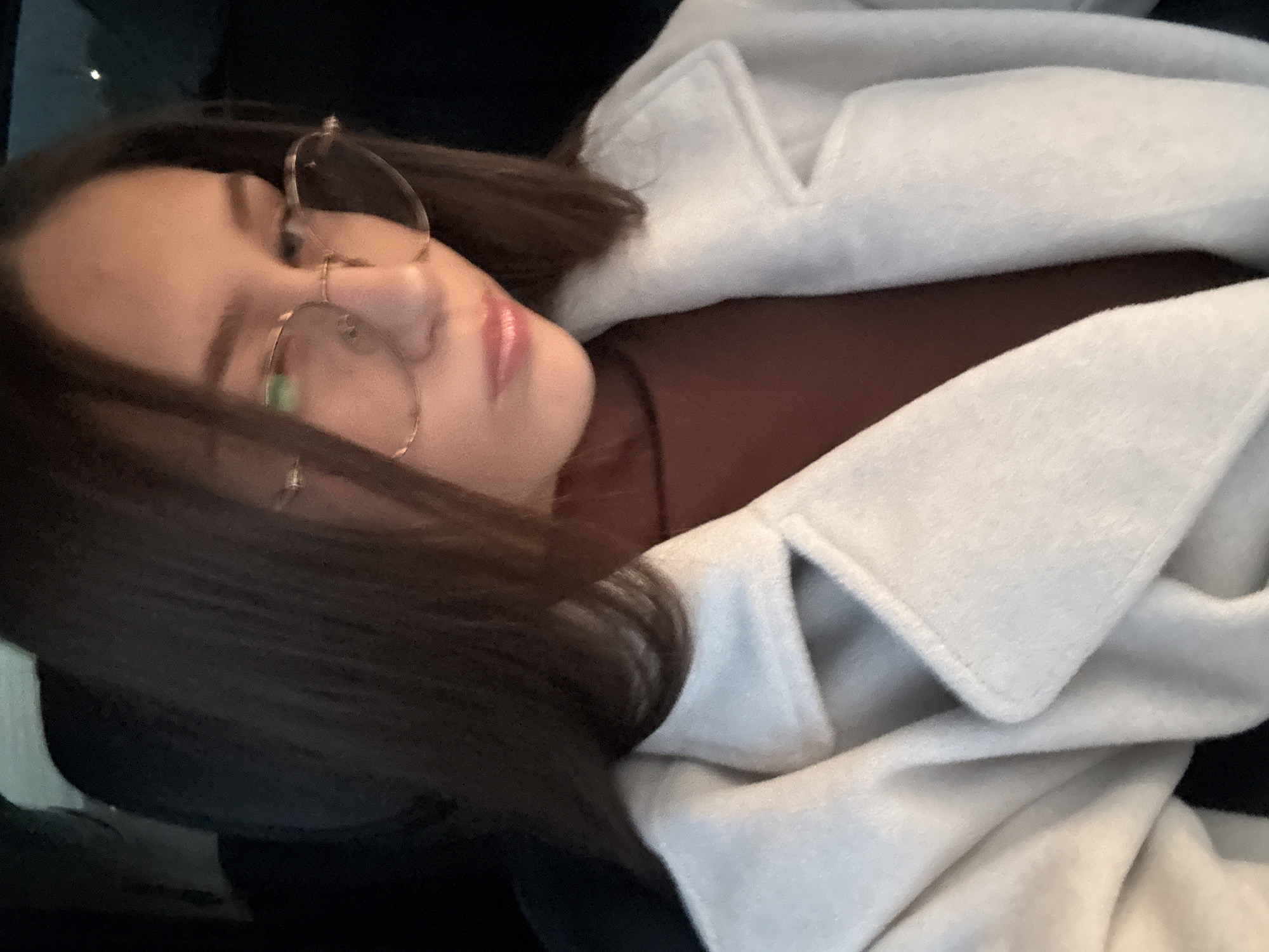 A woman with brown hair and glasses lies down, wearing a brown turtleneck and a white coat. The setting appears to be indoors, potentially in a vehicle. It is not possible to determine the exact location within the city of Sofia, Bulgaria from this image.