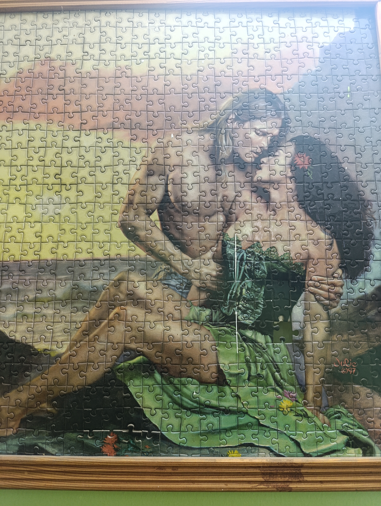 The image shows a framed jigsaw puzzle depicting a romantic scene between a man and a woman. The man is shirtless and has blond hair, while the woman has dark hair and is wearing a green dress. They are embracing, with the man's arm around the woman's shoulder and her hand touching his face. The background appears to be a natural landscape with water and greenery. There is a signature on the puzzle that reads "Julie 1997". The puzzle is placed on a green surface, and the frame is made of wood. The image does not contain any information or clues that would allow for its location to be identified in the city of Moscow, Russia.