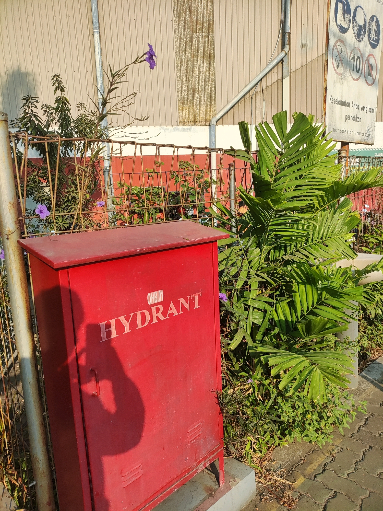 This is a medium shot, eye-level, outdoor, daylight photograph of a red hydrant box in front of a beige corrugated metal building. The hydrant box is on the left side of the frame, angled slightly away from the camera. It has "HYDRANT" and "OHB.01" printed in white capital letters on its front. A shadow of a person holding a camera or phone is cast on the left side of the hydrant box. To the right of the hydrant box are dense green plants, including a large palm with fronds. A rusty metal fence is visible behind the hydrant box and to the left of the plants. In the background, the beige building has a corrugated texture and a silver pipe runs vertically down its facade. A white sign with black circular symbols and Indonesian text is mounted on the building to the right of the pipe. The ground in front of the hydrant and plants is paved with interlocking gray bricks. There are two small purple flowers visible on stalks behind the fence and to the left of the hydrant box. The scene appears to be located in a commercial or industrial area, possibly an office building complex or factory, given the building style and the presence of the hydrant and safety sign. Based on the sign in Indonesian, the location is likely within Indonesia, and specifically East Jakarta, Indonesia, as suggested by the context provided.