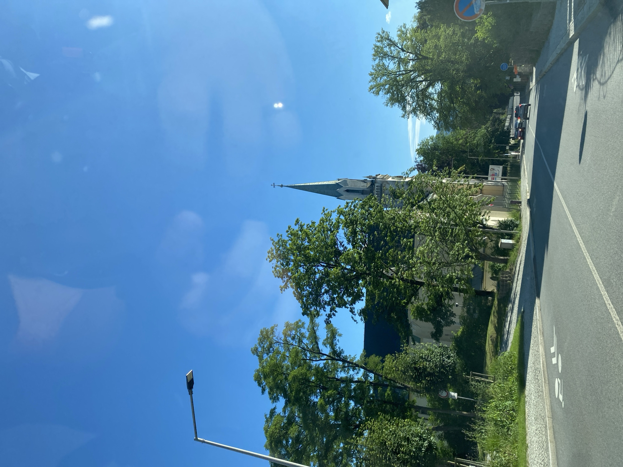 The image depicts a street scene with a church in the background, a street lamp on the left, and trees lining the road. The sky is clear and blue, with some reflections on the surface from which the photo was taken. The road has white markings, including a bicycle lane symbol. There are a few cars on the road. The overall environment suggests an outdoor, urban or suburban setting during the daytime.