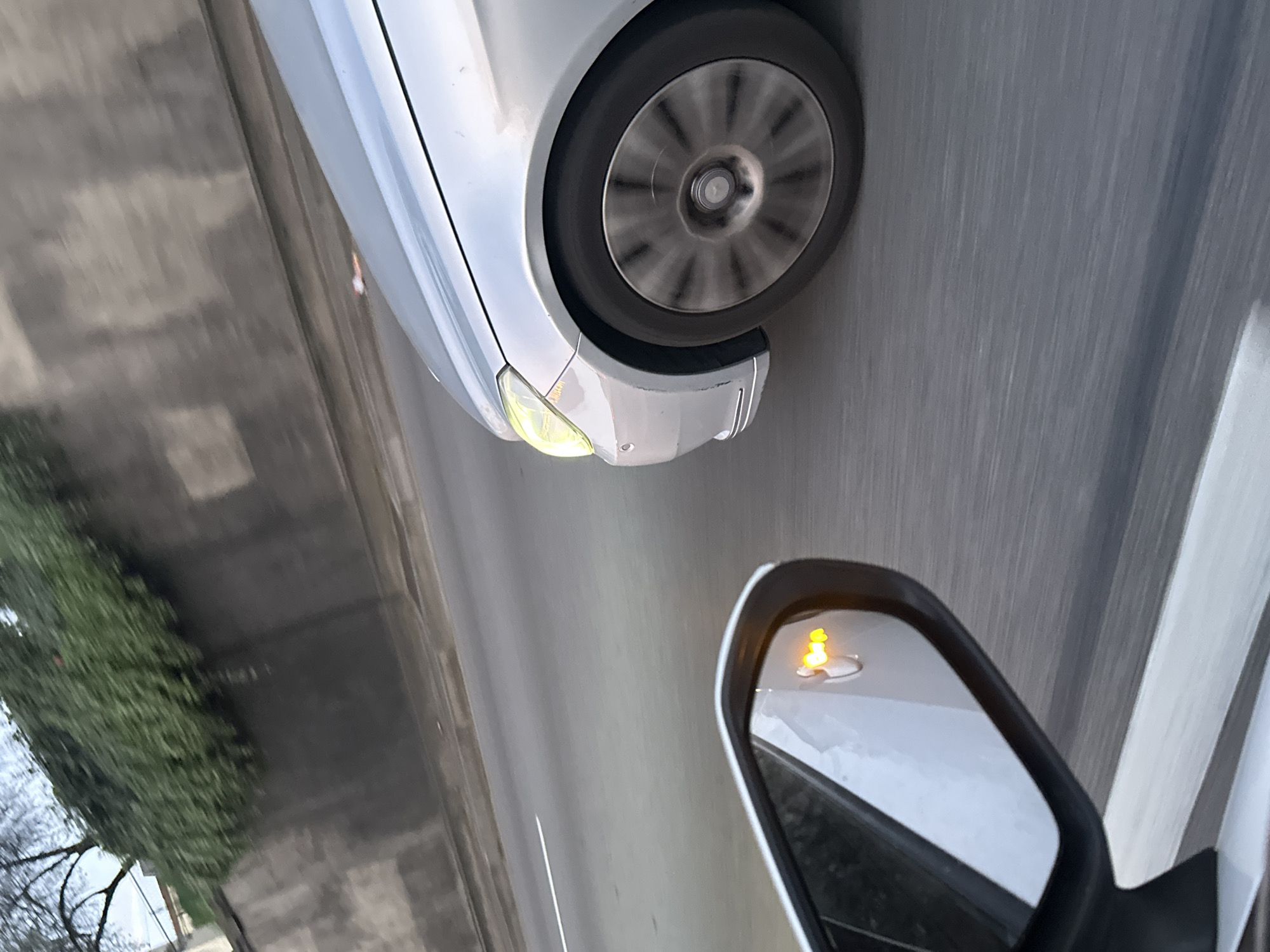 The image is a close-up shot taken from inside a moving vehicle, angled downward toward the road. The bottom right corner shows a side mirror with an active turn signal indicator. To the left of the mirror is the asphalt road with white lane markings. Further left, the image shows the front portion of another silver car, including its front wheel and turning indicator. In the background, out the window, is a row of trees and a glimpse of buildings. The lack of landmarks and the general nature of the scene prevent identifying Bagnolet, France as the definite location with the provided data.