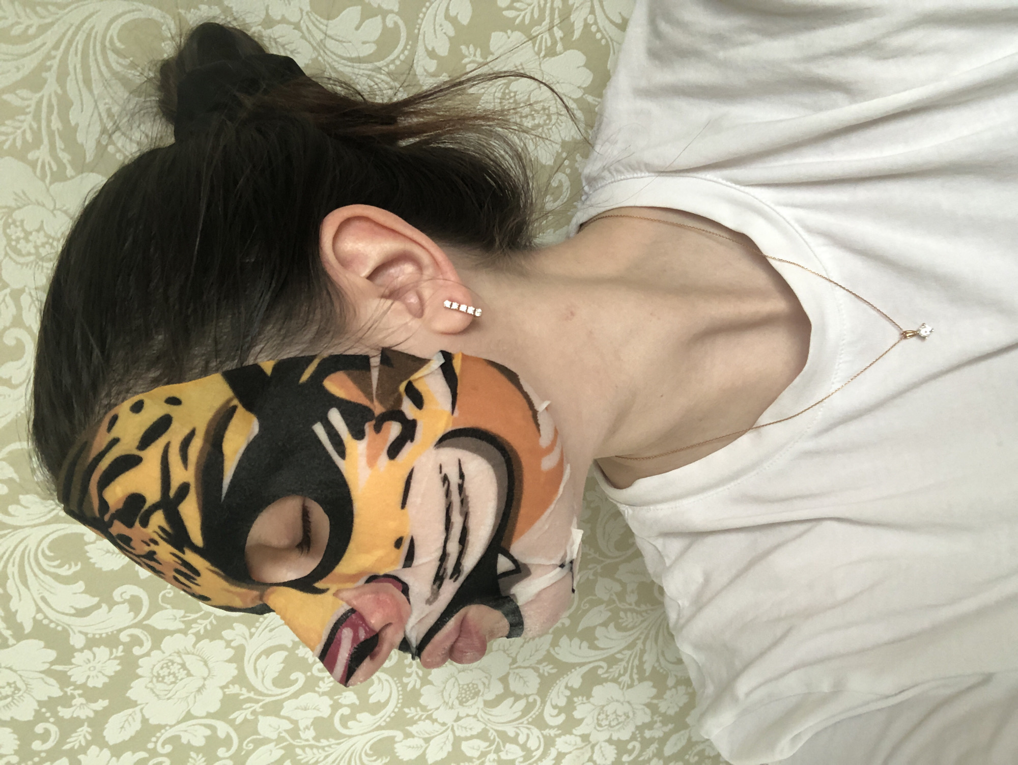 A person lies on a patterned fabric surface, possibly a bed or couch, wearing a white t-shirt and a patterned face mask. The face mask is predominantly orange and black, with additional colors including pink and white. The person's dark hair is pulled up into a bun, and a gold necklace is visible on their chest. A small earring adorns their ear. The patterned fabric beneath the person has a floral design with light and dark tones of green. The location is not apparent from the image.