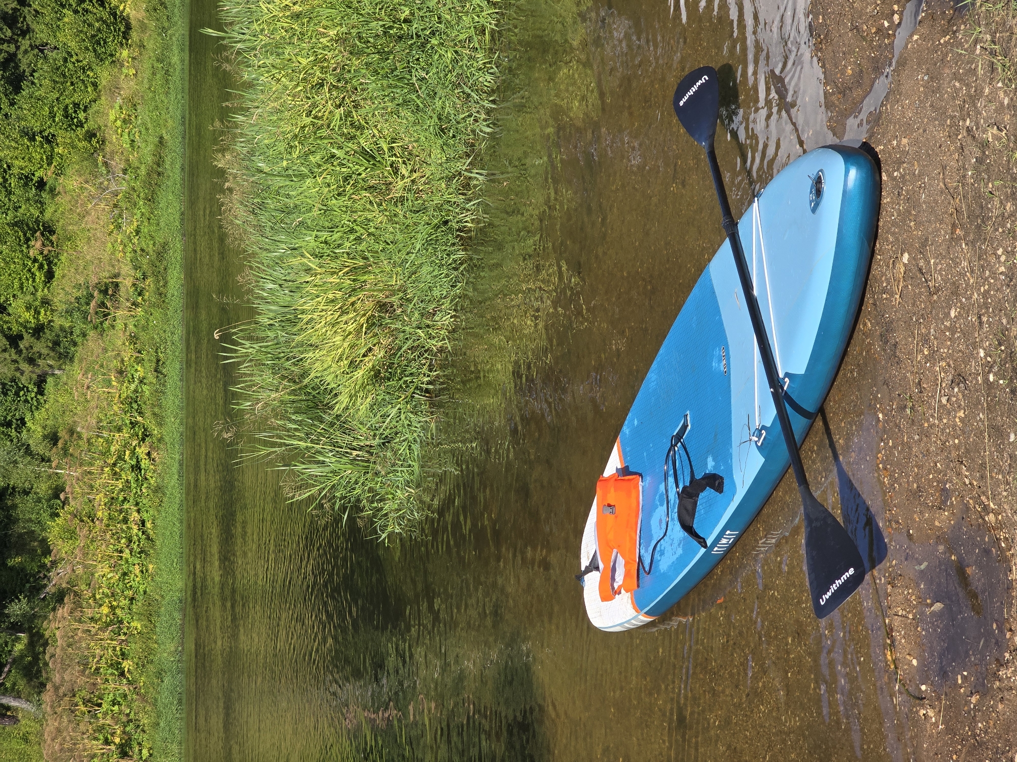 A stand-up paddleboard with an orange life vest and a paddle lies partially in the shallow water of a river, with its front end on the gravelly shore. The river is surrounded by lush green vegetation and trees, suggesting a natural, unpopulated area. The water reflects the sky and surrounding foliage. There are no people visible in the image. The location is the city of Voinskaya Chast' Mchs, Russia.