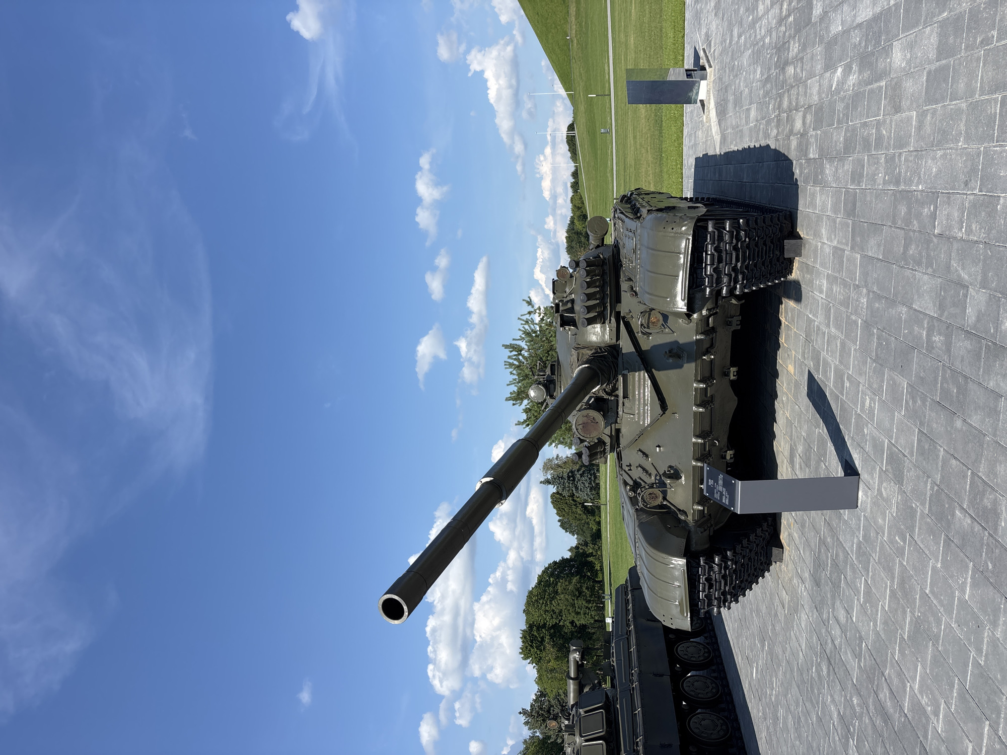 The image captures a frontal view of a military tank displayed on a paved area under a partly cloudy blue sky. The tank, painted olive drab, is positioned prominently in the foreground with its long gun barrel pointing upwards and to the left. Its heavy tracks and robust chassis are clearly visible. To the left of the main tank, a portion of another similar tank is visible, suggesting a military exhibit. In the background, a green, grassy landscape stretches out, featuring a gentle hill on the right side of the frame. The environment appears to be an outdoor museum or memorial site, possibly located in the city of Ozyaritska-Slabadski rural council, Belarus, given the context. There are no people visible in the image.